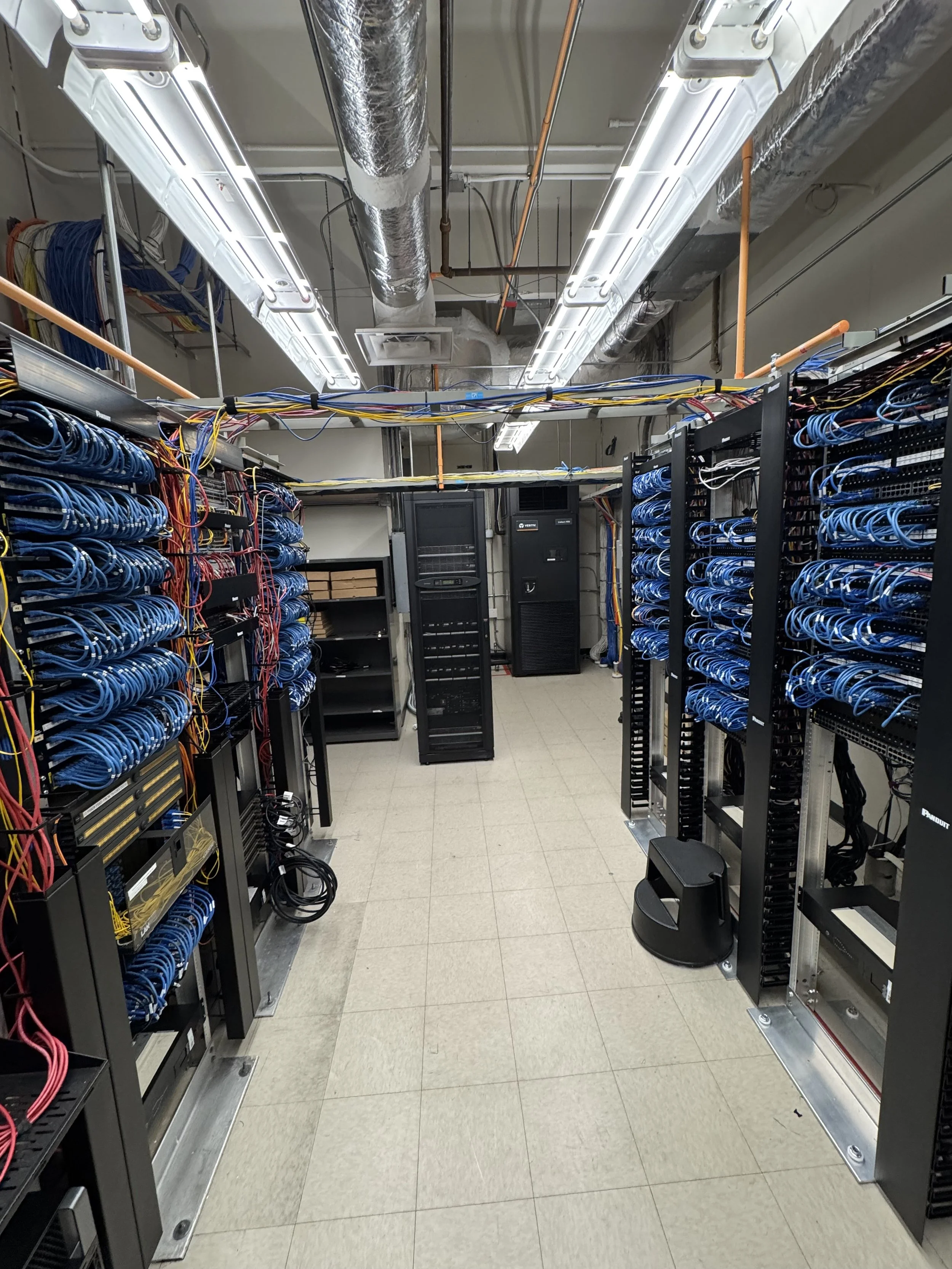 A server room with multiple server racks filled with blue networking cables, and a black server cabinet in the center. There are bright fluorescent lights on the ceiling and visible ventilation ducts.