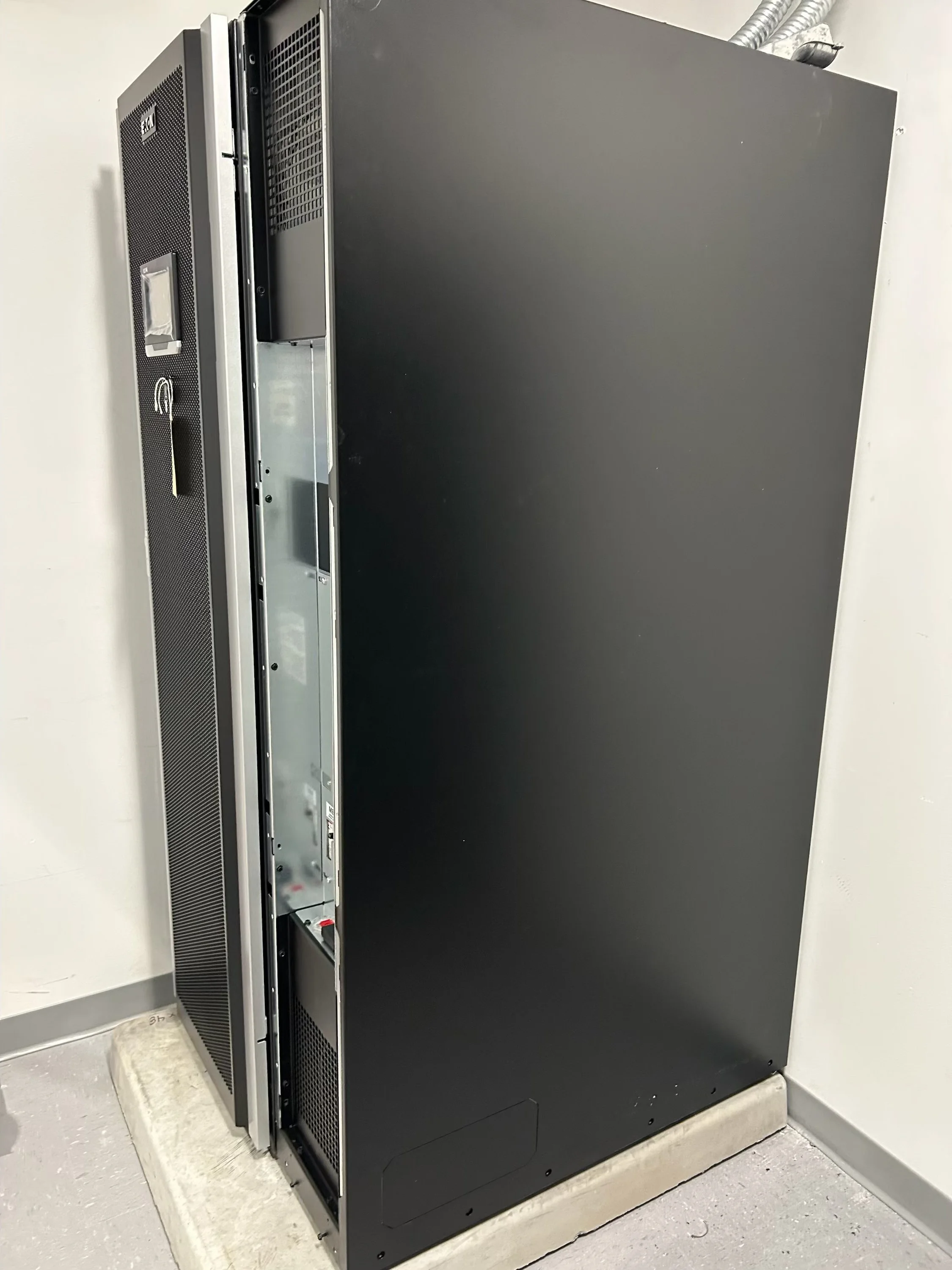 A large electronic equipment cabinet or server rack with a black exterior, positioned on a concrete floor against a white wall. It has ventilation slots and some internal components visible through the open front panel.