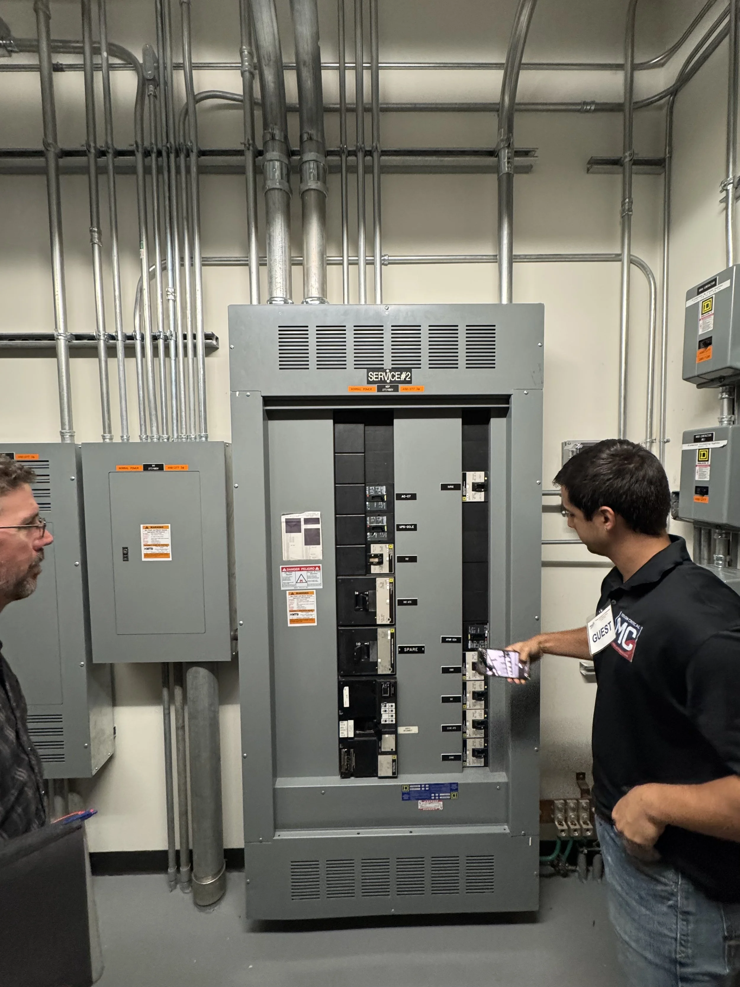 Two men standing in front of an electrical panel and circuit breakers, engaged in conversation, in an industrial or utility room.