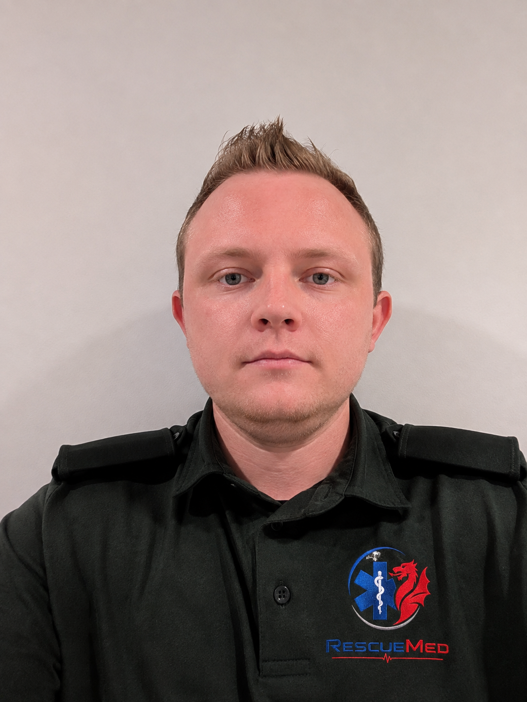 A man with short, styled blond hair wearing a black RescueMed uniform shirt with a logo of a red dragon, blue medical symbol, and red heartbeat line on the chest, standing against a plain light-colored background.