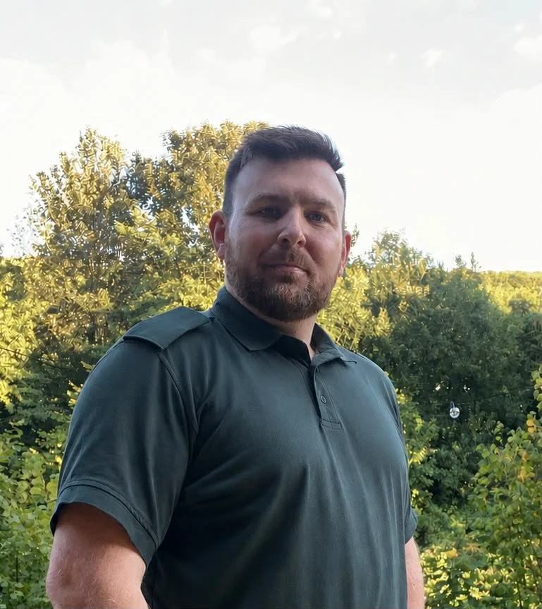 Man with short brown hair and beard standing outdoors among trees, wearing a dark green polo shirt.