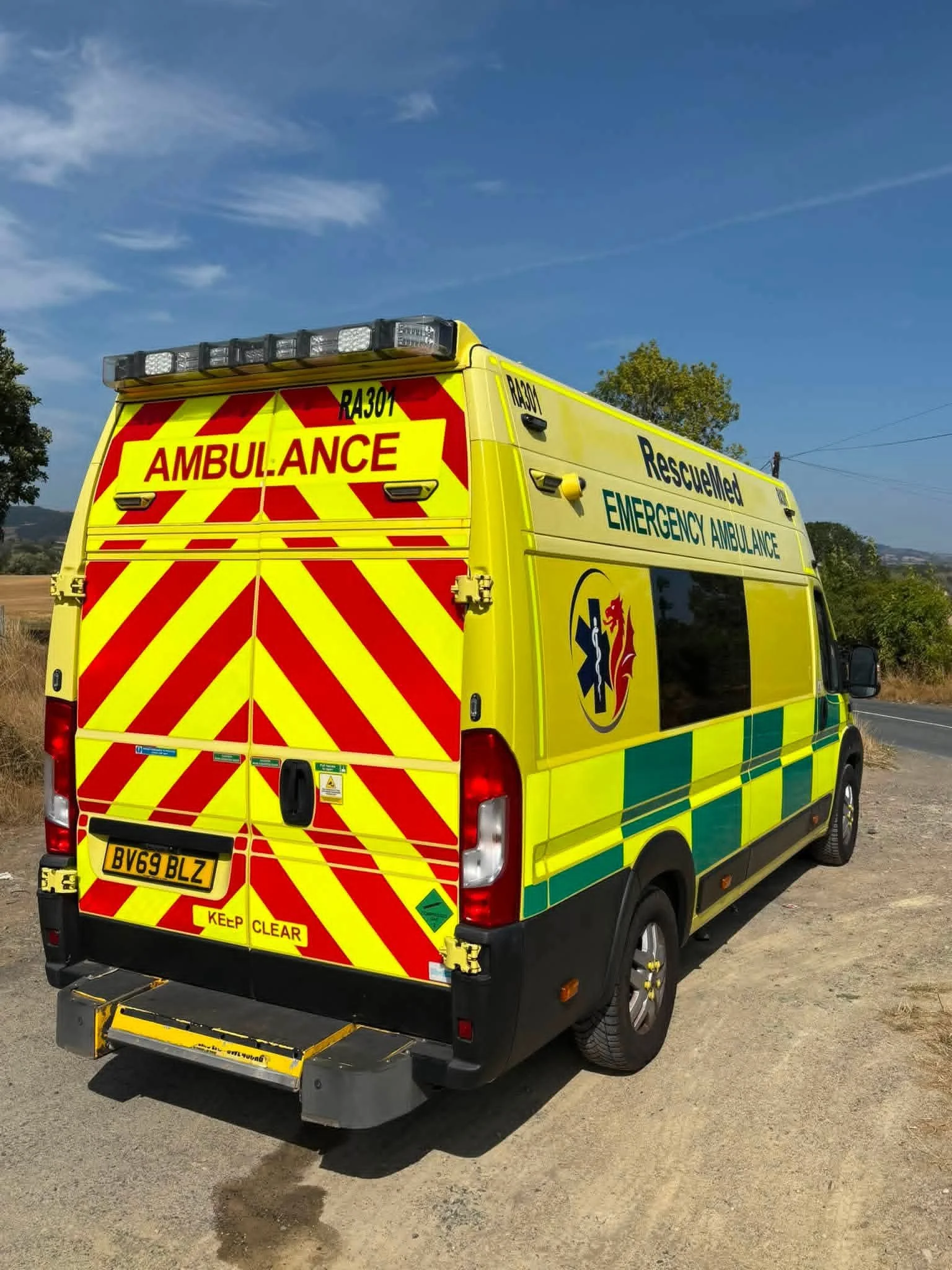 Yellow emergency rescue ambulance with red and yellow chevron pattern on the back doors, parked on a dirt road with trees and hills in the background under a blue sky.