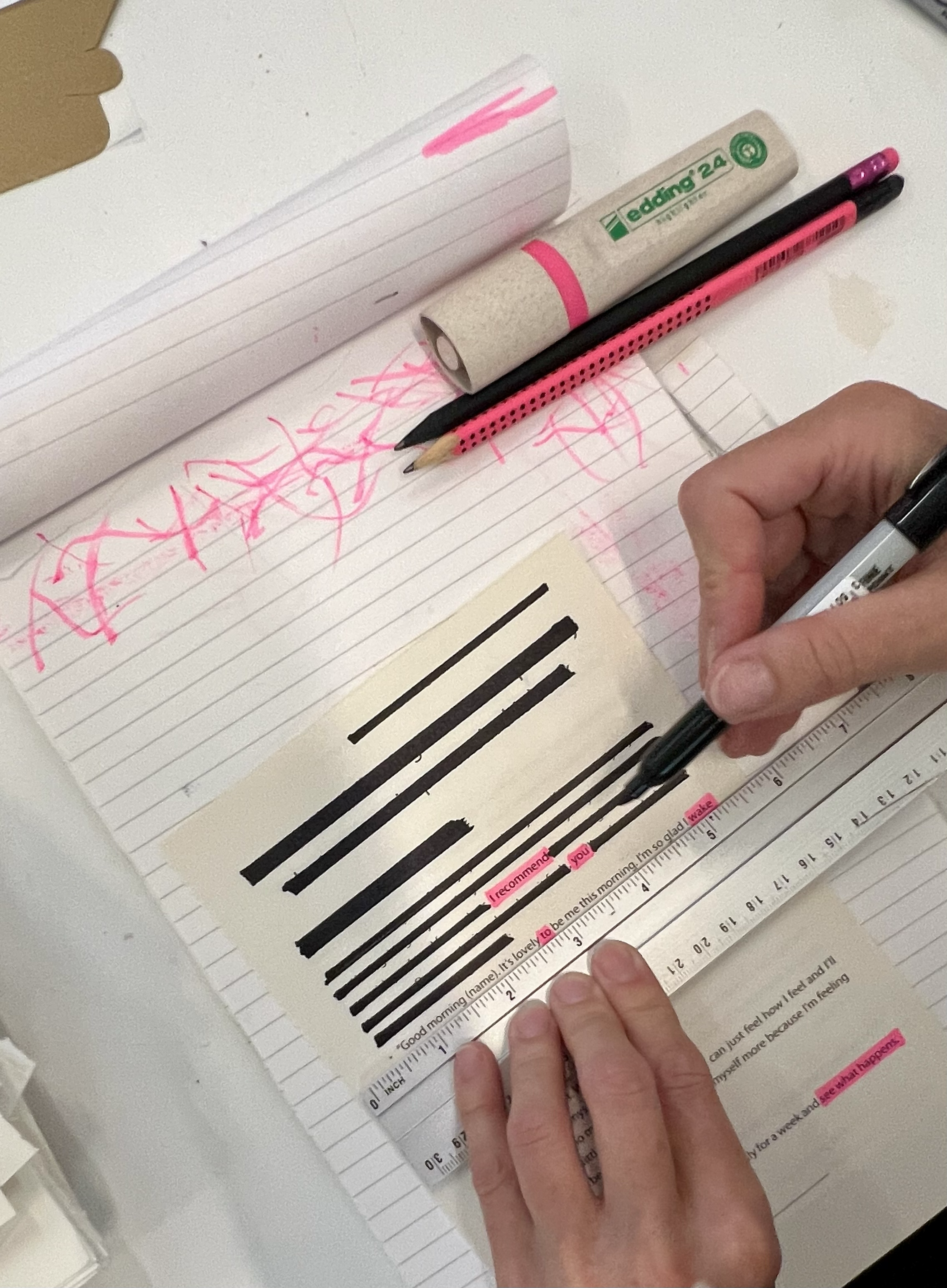 A person writing in a notebook with a black pen, with pink highlights on the pages. There is a ruler and highlighter on the table along with a piece of paper with pink highlighted text.