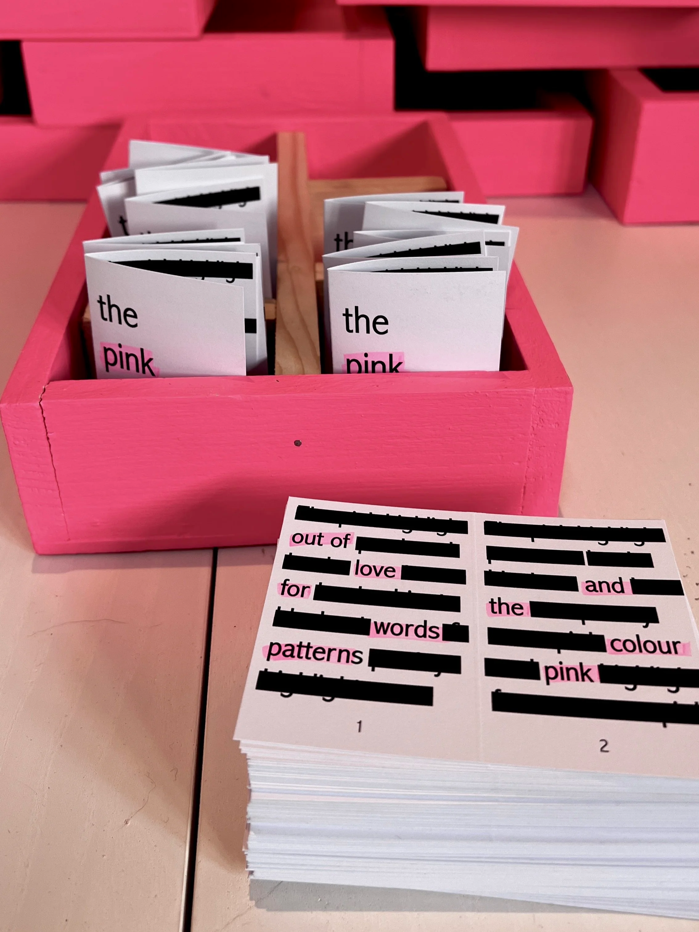 Pink wooden tray holding small booklets with words 'the' and 'pink' on the covers, and a printed sheet with words 'out,' 'love,' 'for,' 'words,' 'patterns,' 'colour,' and 'pink,' some blacked out.