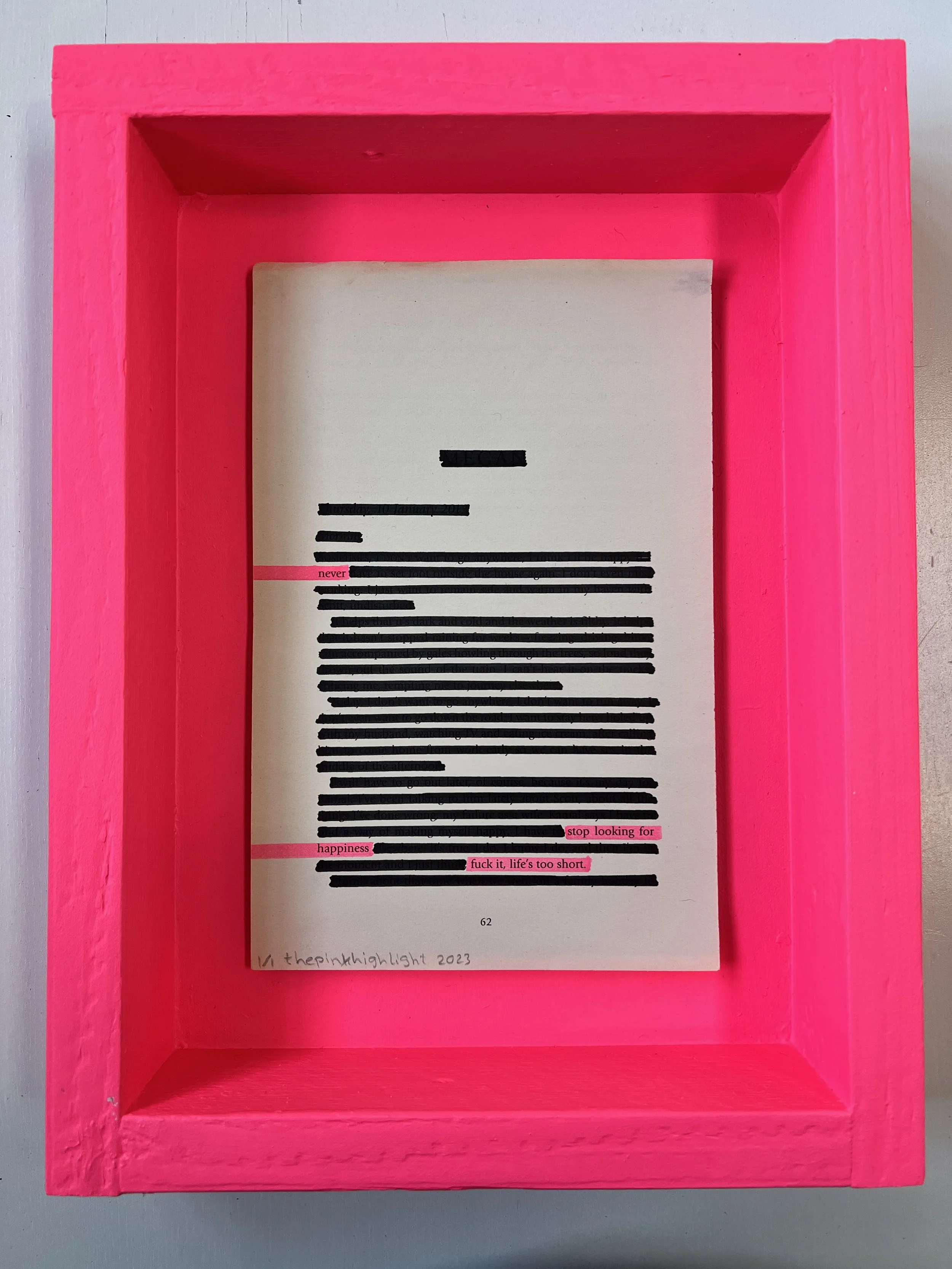 A book open to a page with text blacked out, except for a few words highlighted in pink and a handwritten note at the bottom reading "1/ thepinlight 2023." The book is inside a bright pink wooden frame, placed on a gray surface.