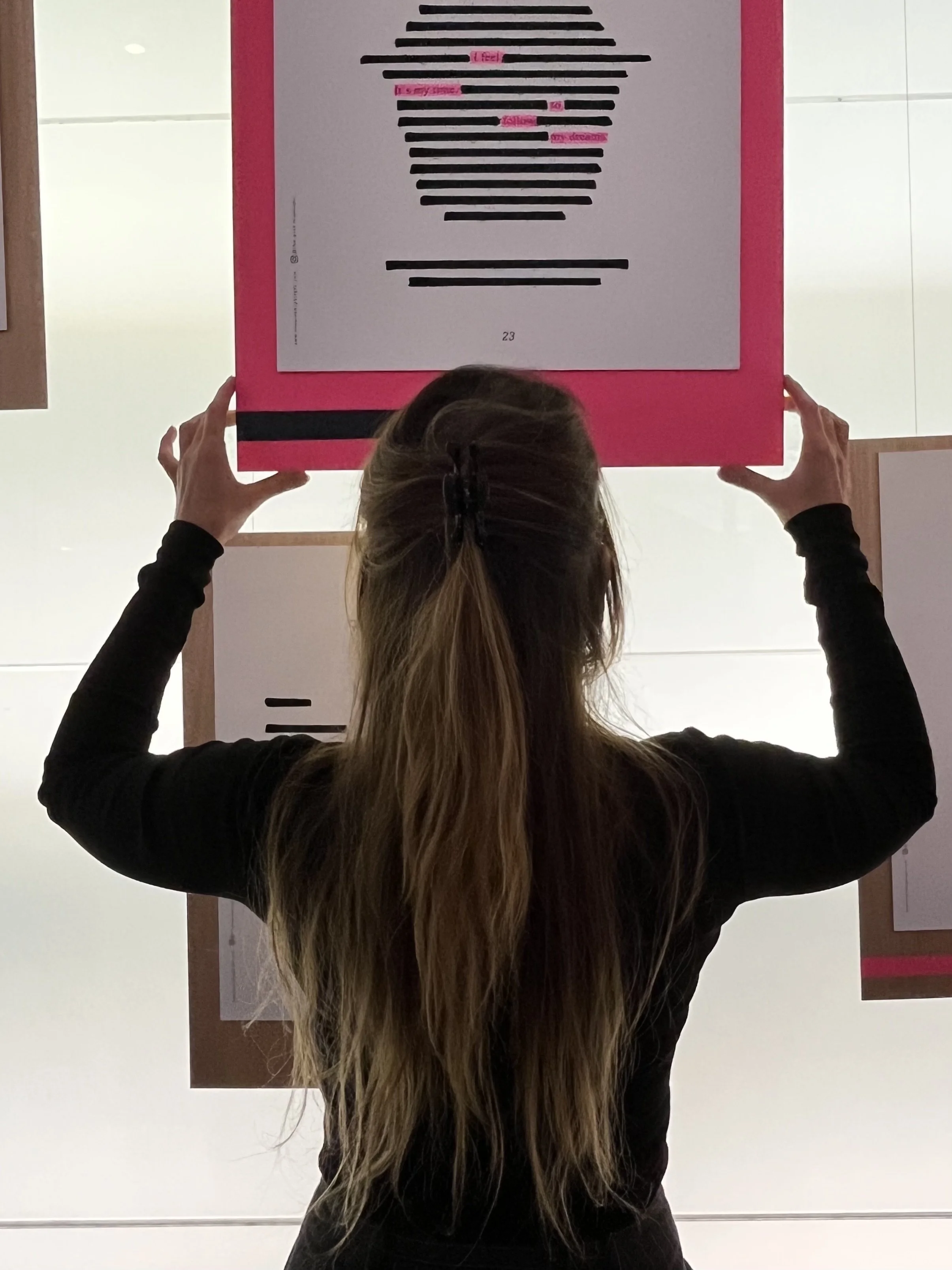 A person with long hair seen from behind holding a square framed artwork against a white wall. The artwork features black horizontal bars creating an abstract shape, with pink text labels on some bars.