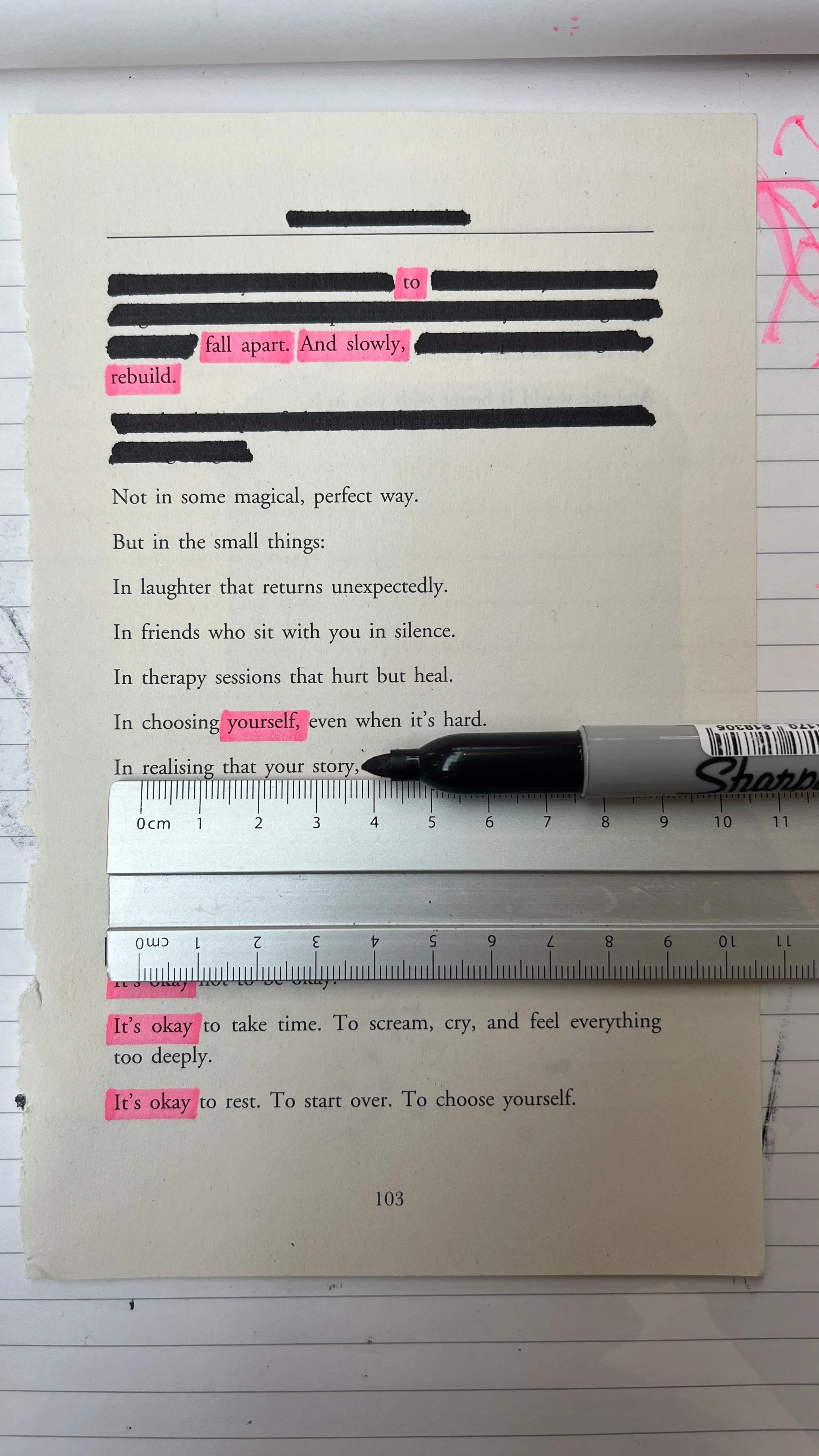 A printed page with text highlighted in pink, showing phrases like 'fall apart,' 'And slowly,' 'rebuild,' 'yourself,' and 'It's okay.' There are black marker strike-throughs on some lines, a Sharpie marker, and measuring rulers placed on the page.