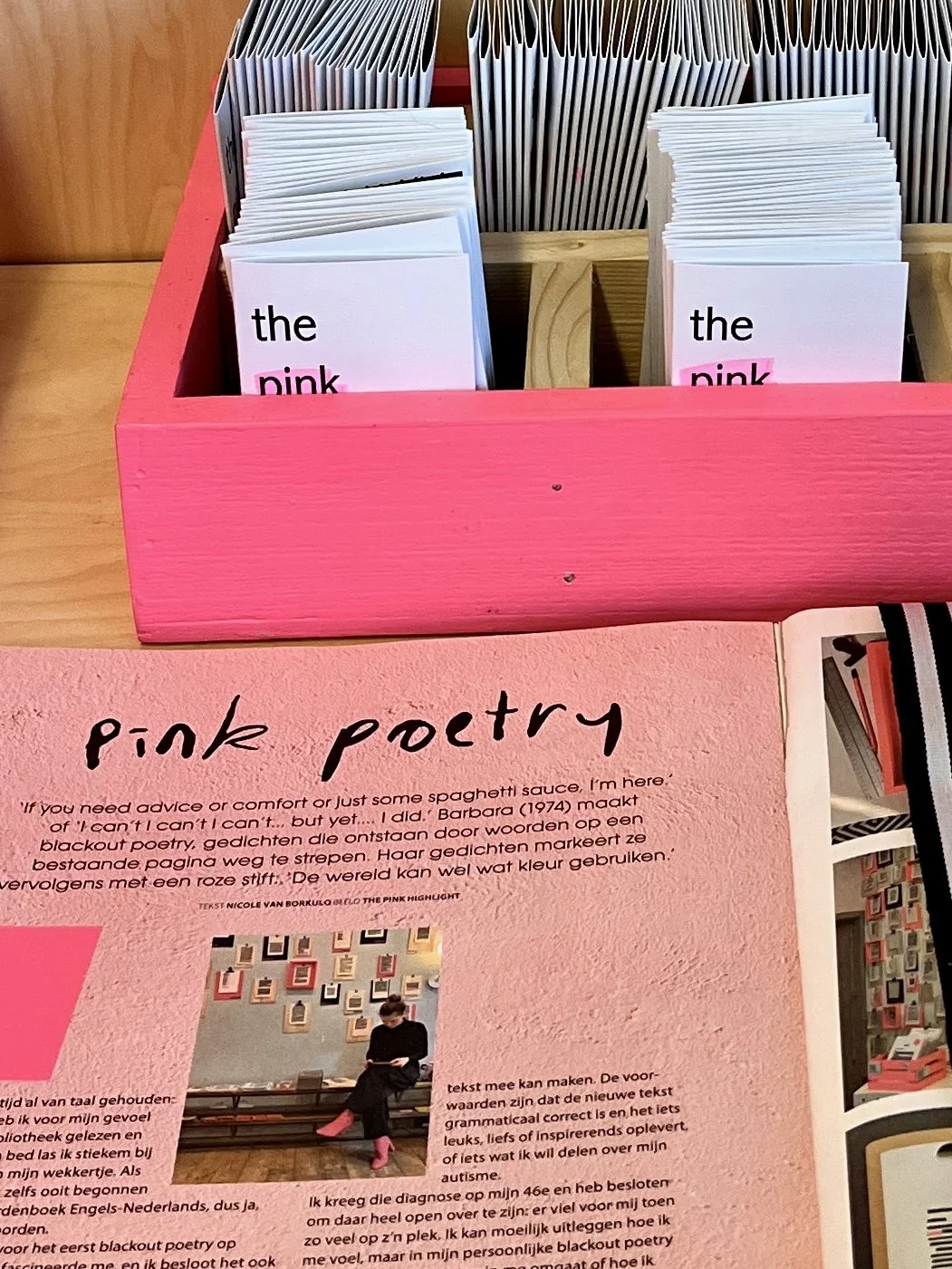 Pink poetry book titled "Pink Poetry" with text and a photo on the page, surrounded by pink and white pocket-sized books labeled "the pink" in a pink and natural wood display box.