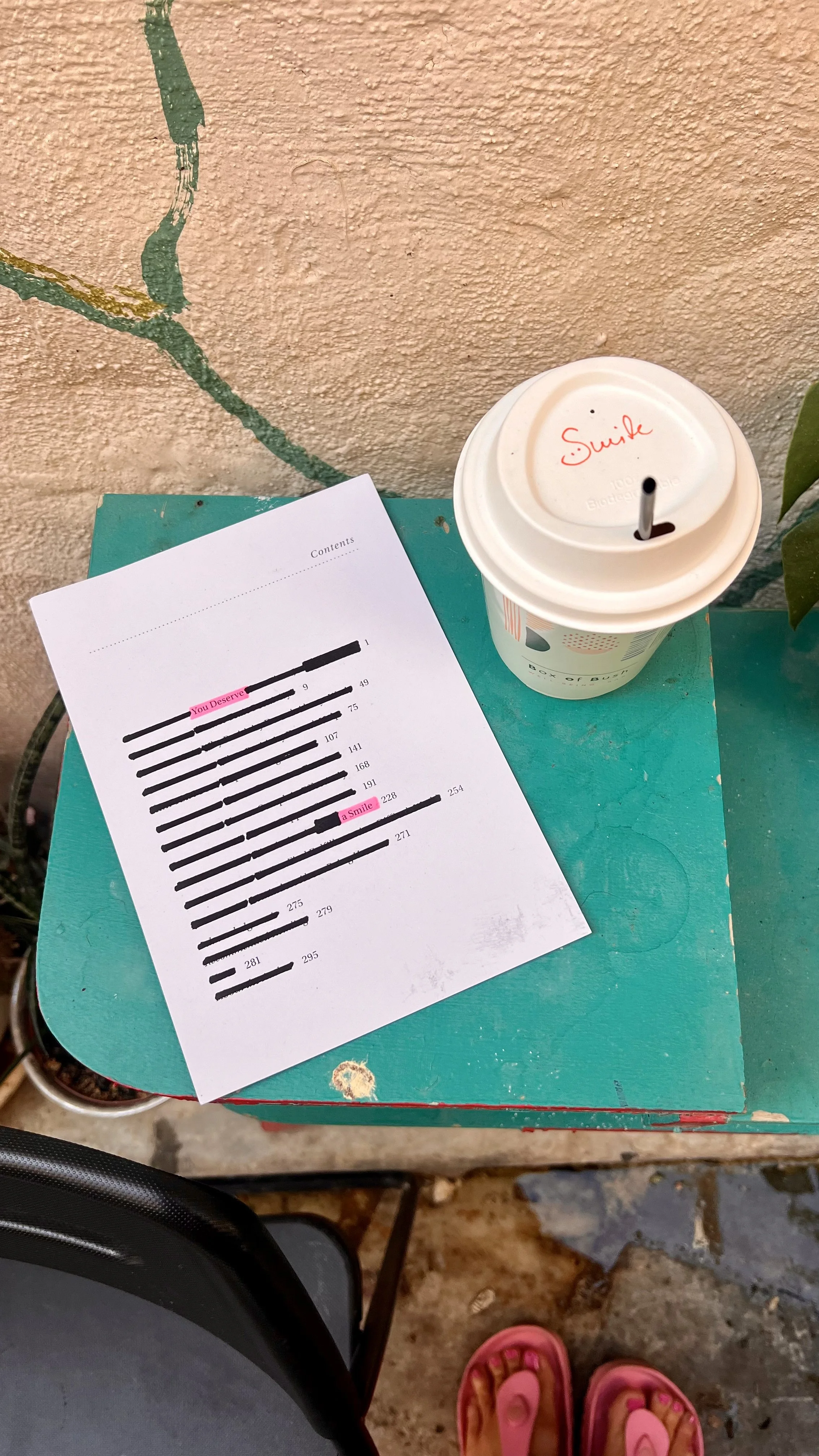 A table with a piece of paper showing a barcode with some text and two pink-highlighted phrases, a disposable cup with a straw labeled 'Smile', a plant partially visible, and a person's pink sandals and grey pants.