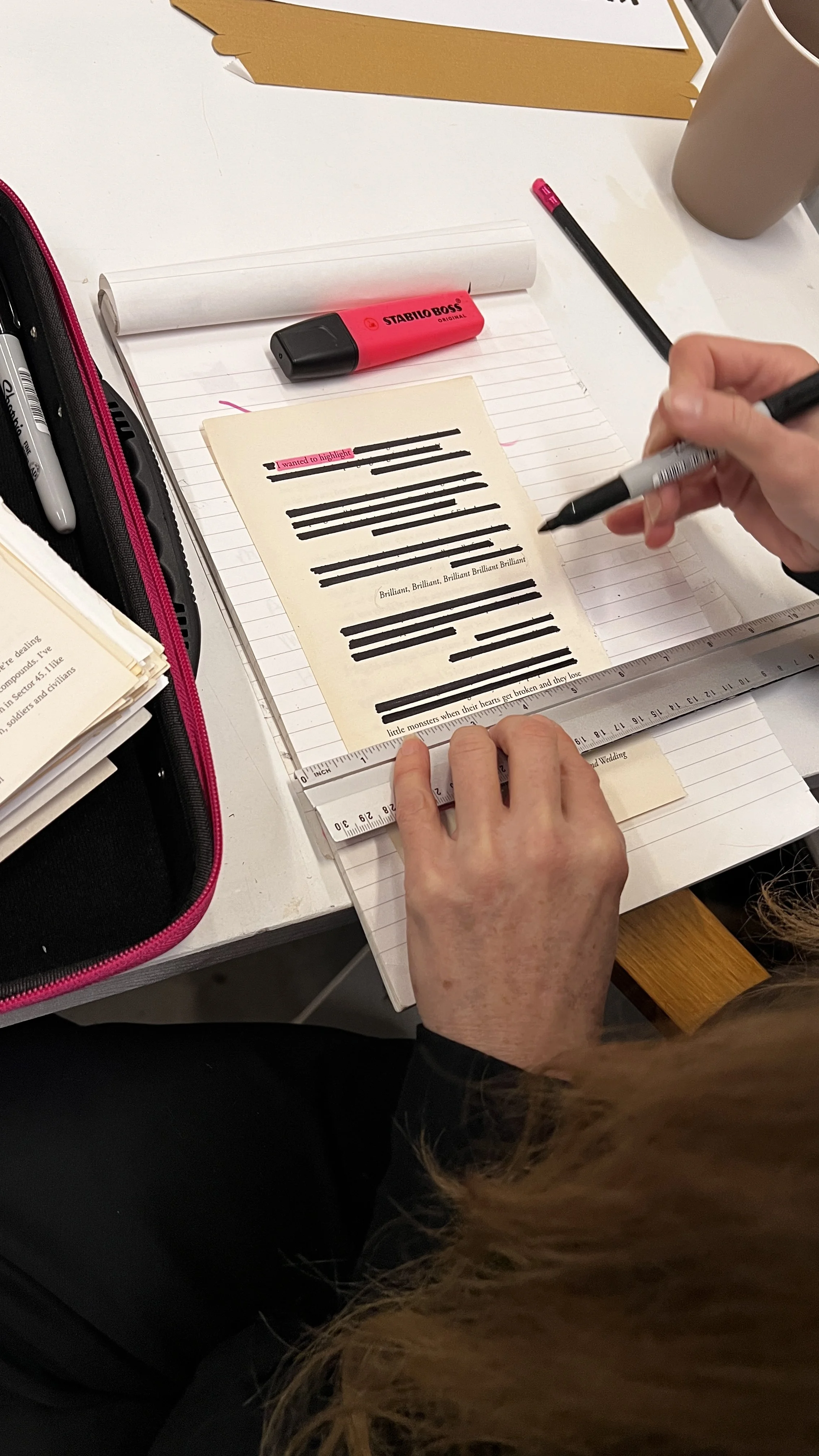 Person using a black marker to highlight a printed document with the word 'Brilliant' repeatedly printed on it, on a desk with a ruler, a pink and gray highlighter, a cup, and some documents.