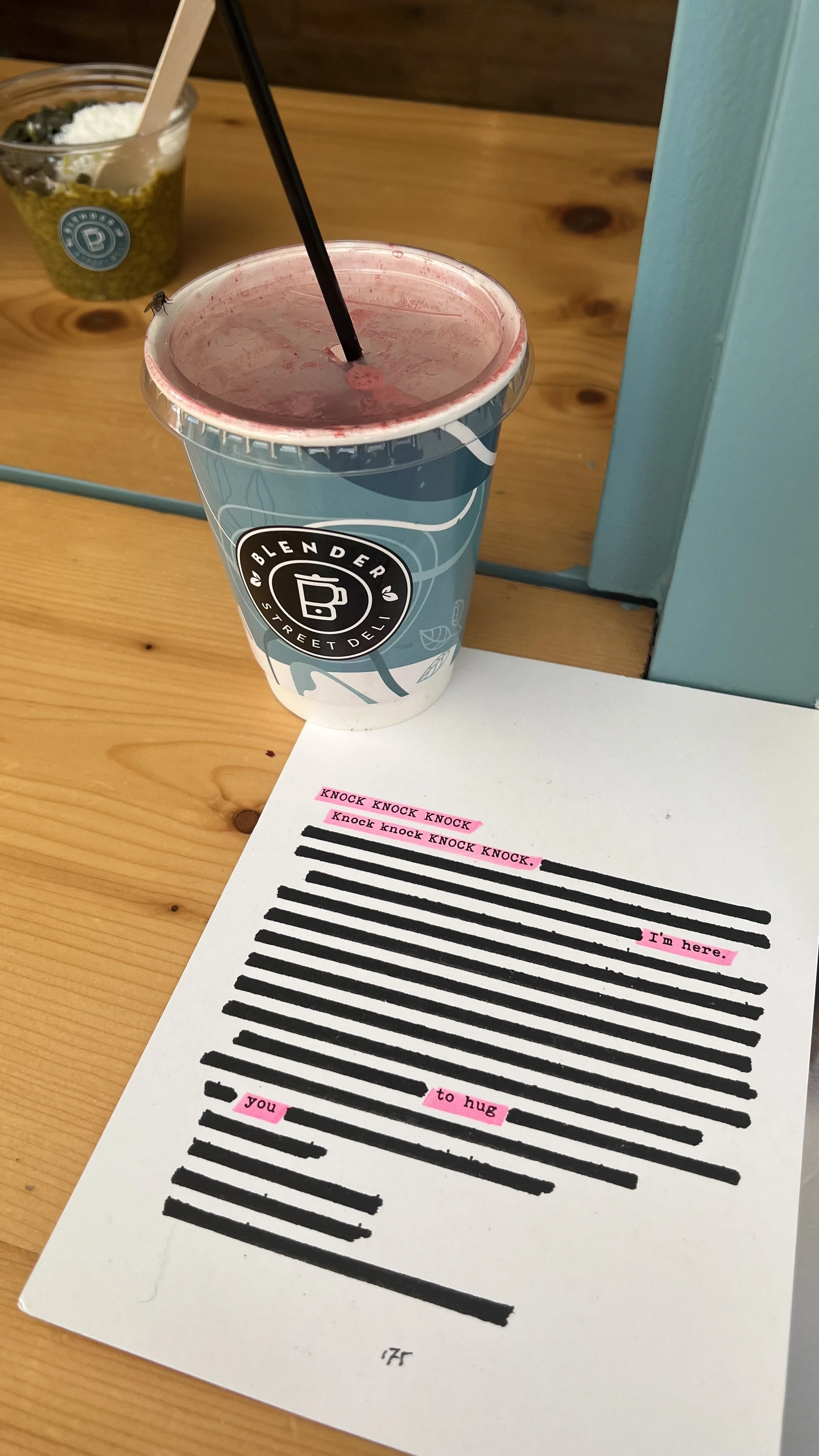 A paper with lines and highlighted words forming a message, a to-go cup with a straw, and a small container of food with a spoon in the background on a wooden table.