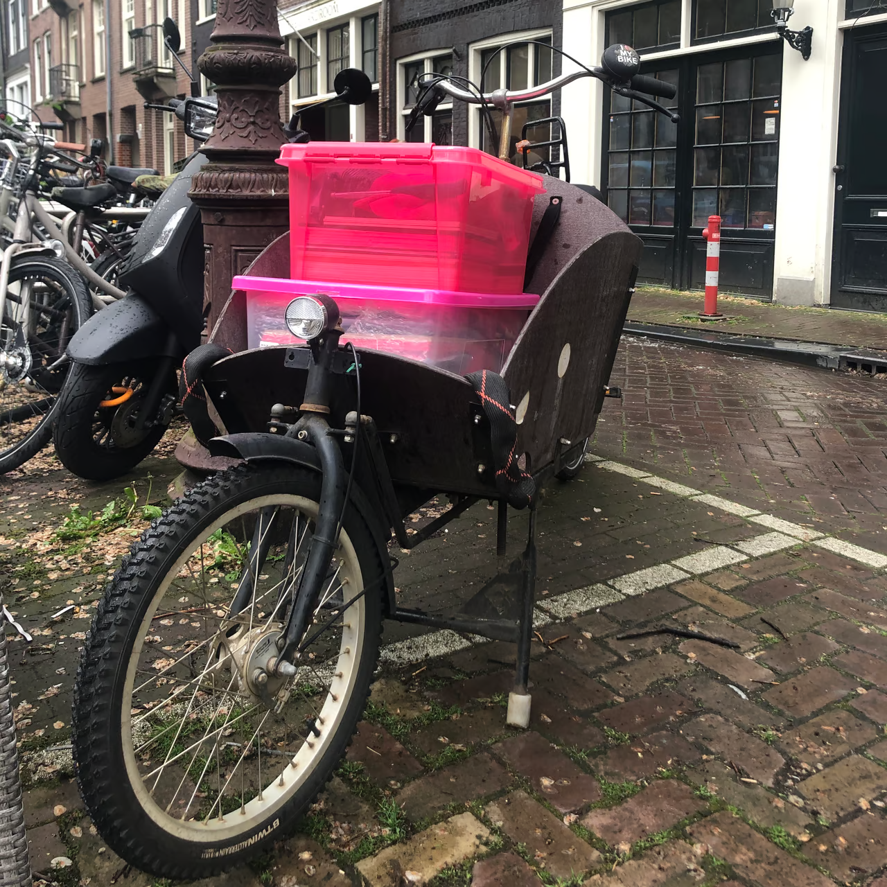 A cargo bike parked on a brick street with pink plastic storage containers in the front compartment. Other bicycles are visible in the background.