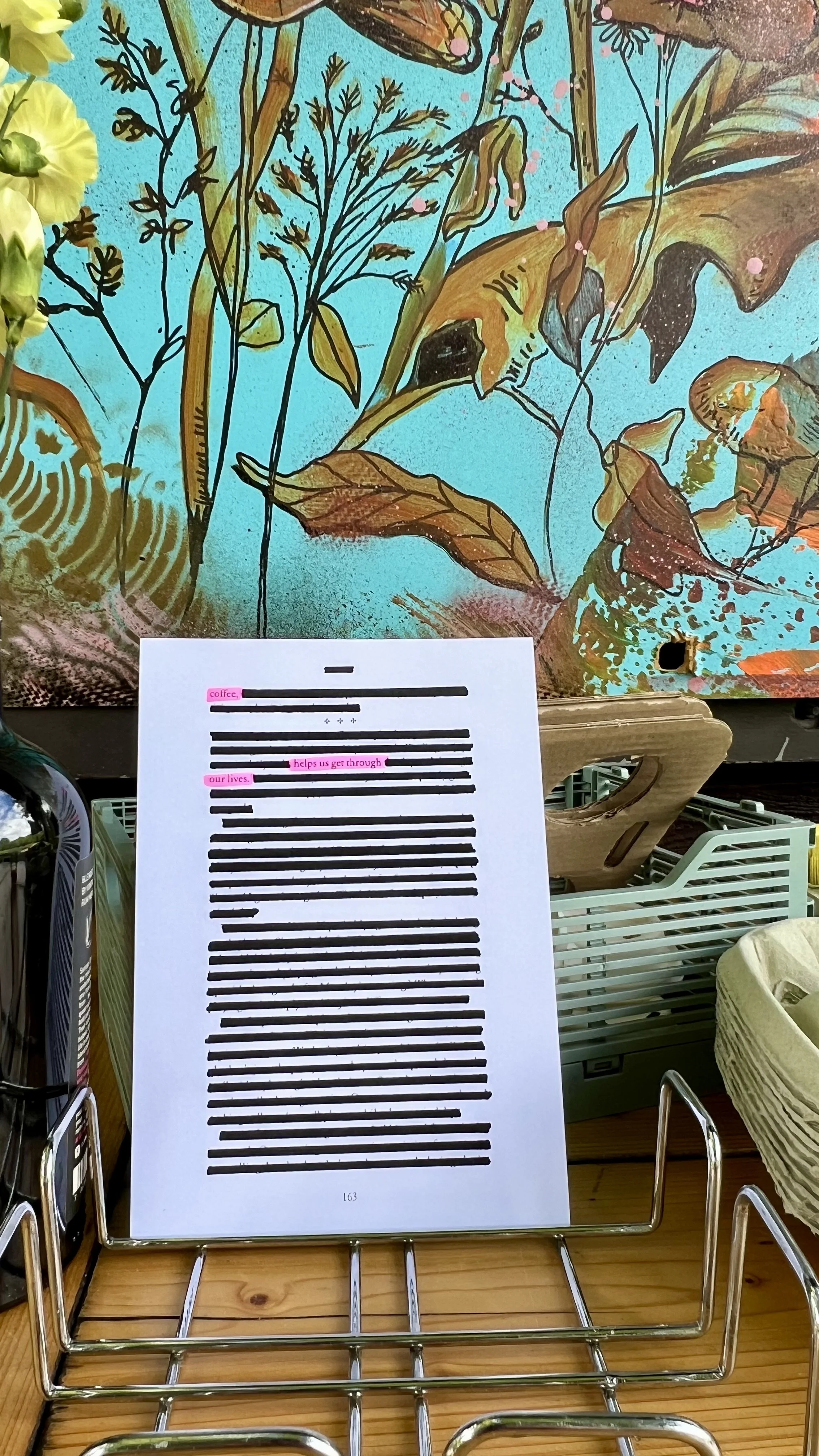 A clipboard with blacked-out lines of text, highlighting the phrase 'coffee helps us get through our lives' in pink, is placed on a wooden table. Behind it, a colorful painting of plants with a bright blue background is visible.