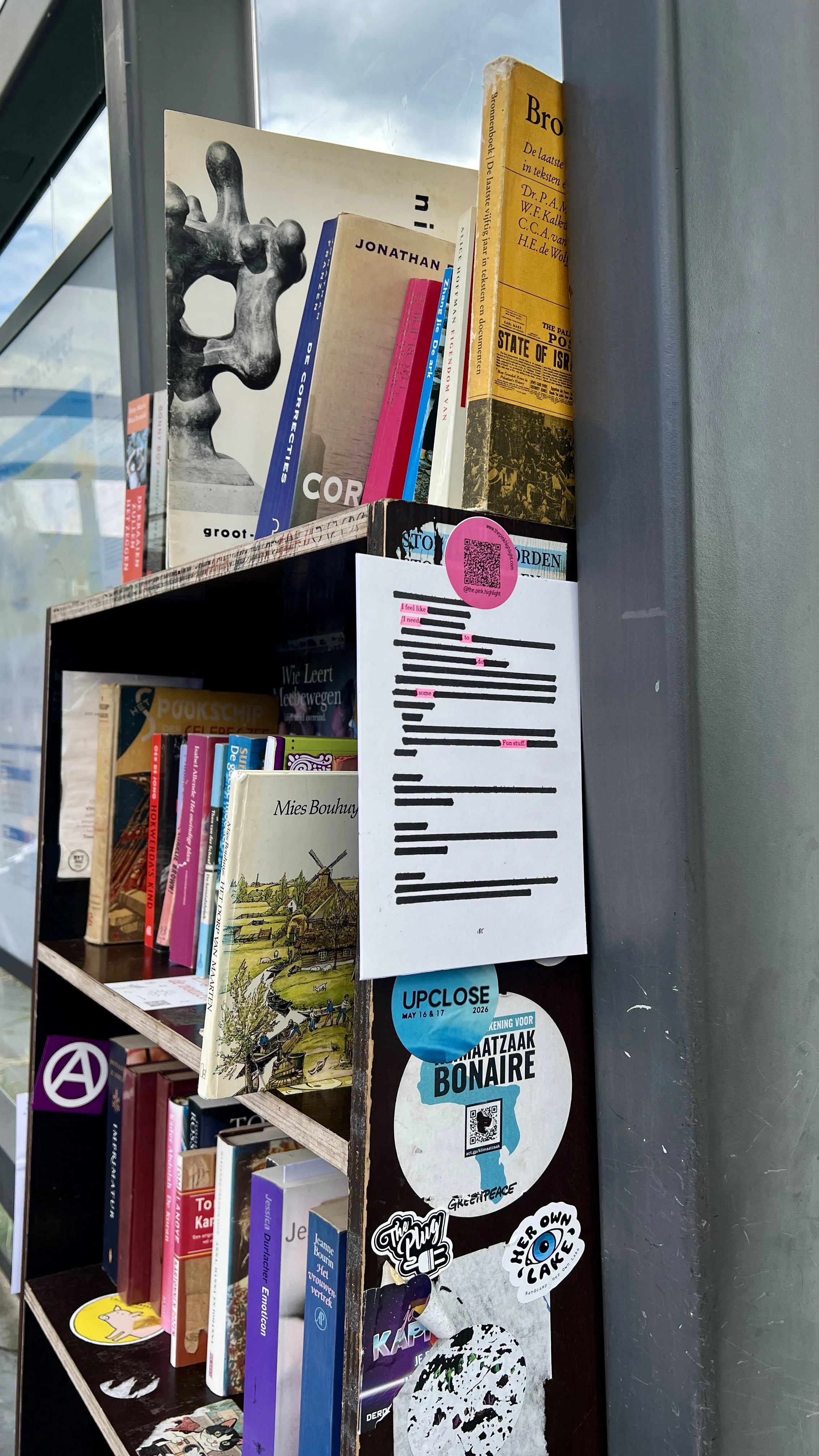 Books stacked on a bookshelf with various posters and stickers attached to the side.