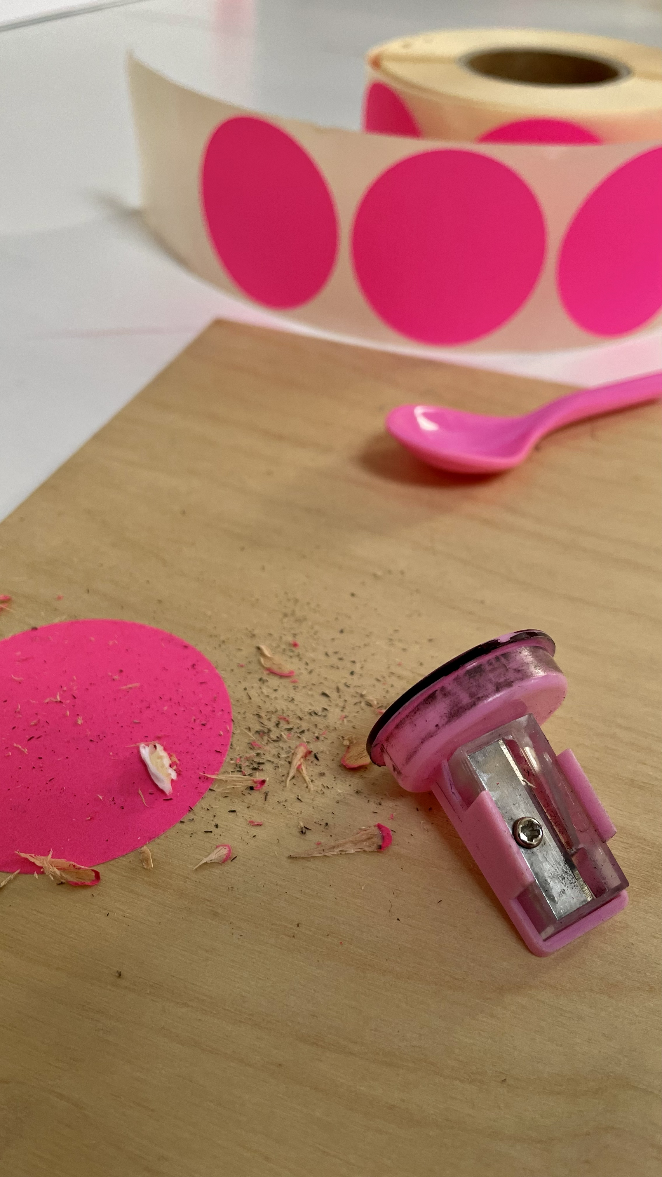 A pink craft hole puncher lying on a wooden surface, with pink paper circles, shavings, and a roll of pink polka-dotted paper in the background.
