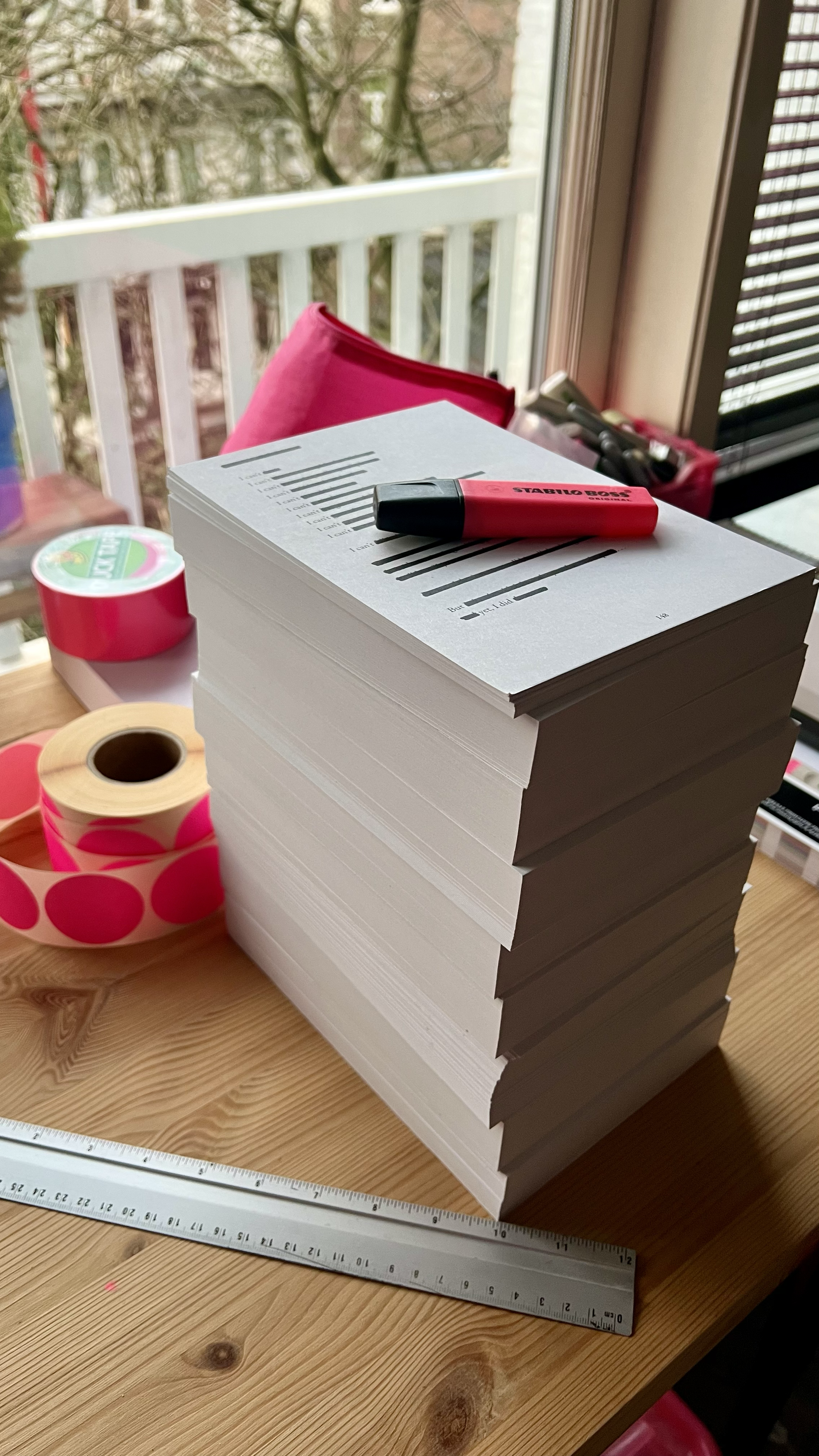 A large stack of white papers or possibly envelopes on a wooden table, with markers, decorative tape, a ruler, and a pink highlighter on top. There is a pink folder in the background, a window with a view of trees, and various office supplies.