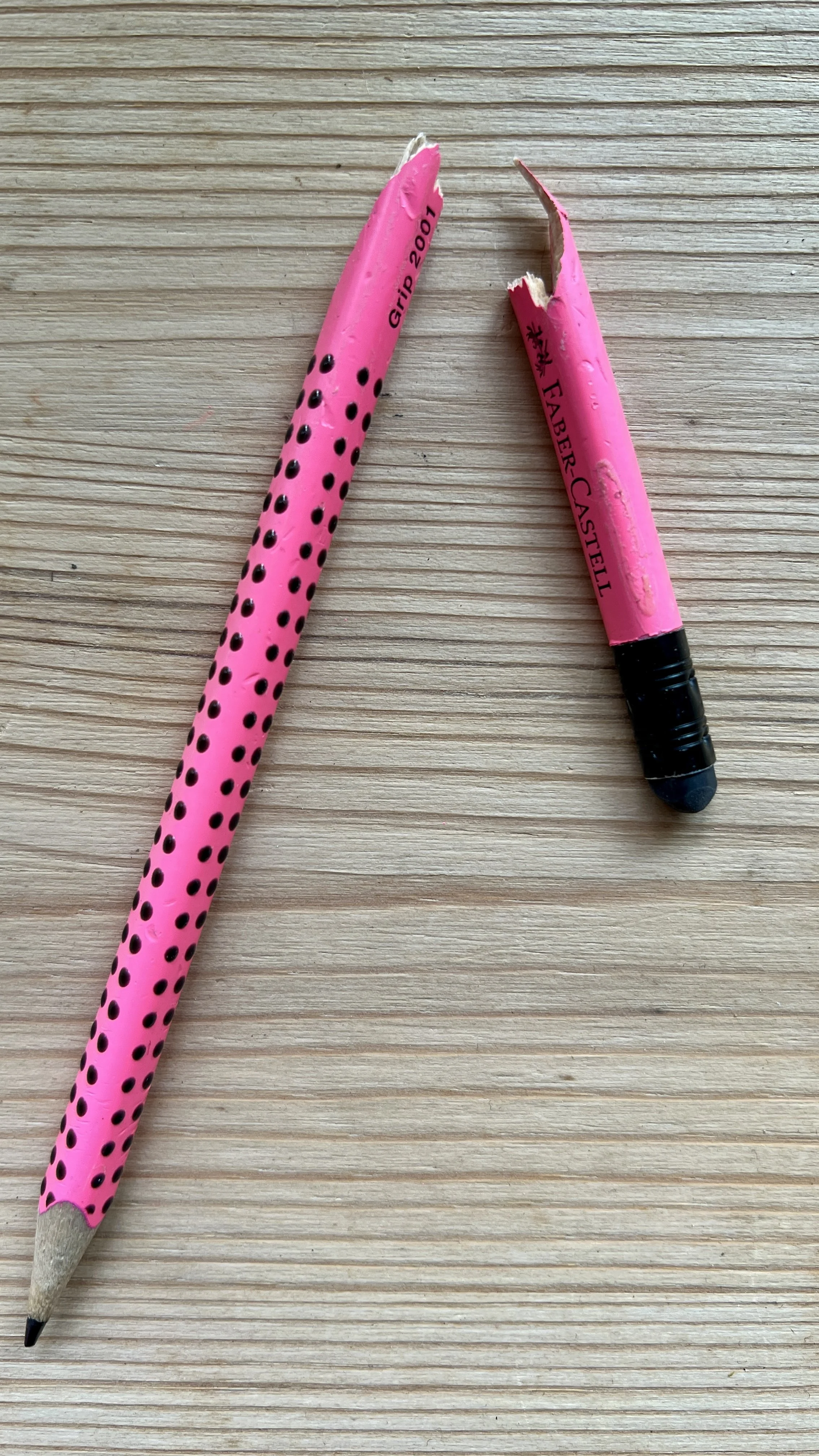 A broken pink polka dot pencil and a pink pen with a black tip on a wooden surface.