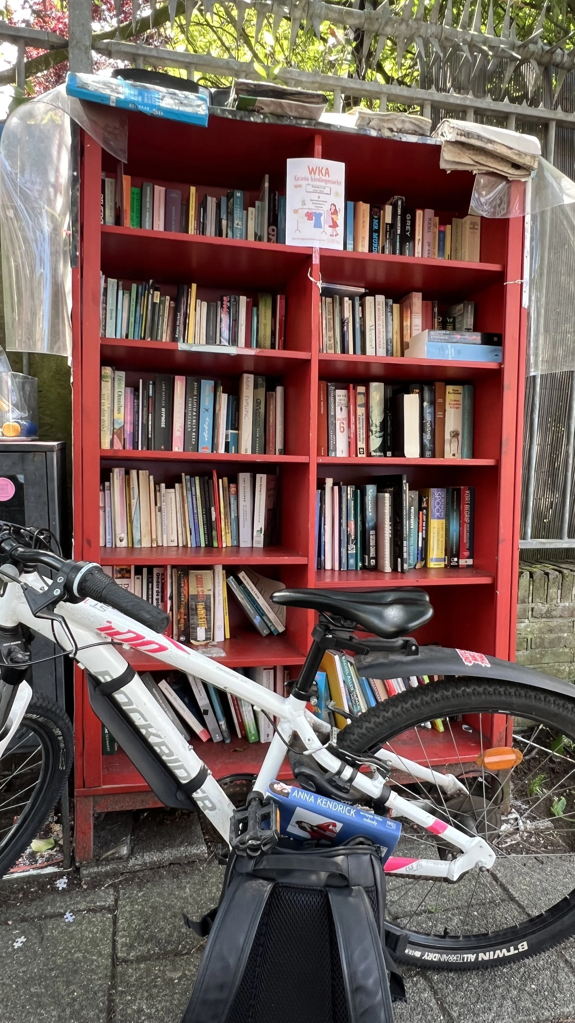 A large red bookcase filled with books situated outdoors behind a bicycle. The bookcase is covered with some transparent plastic sheeting on top. A bicycle is in the foreground, partially blocking the view of the bookcase. There is a small sign on to