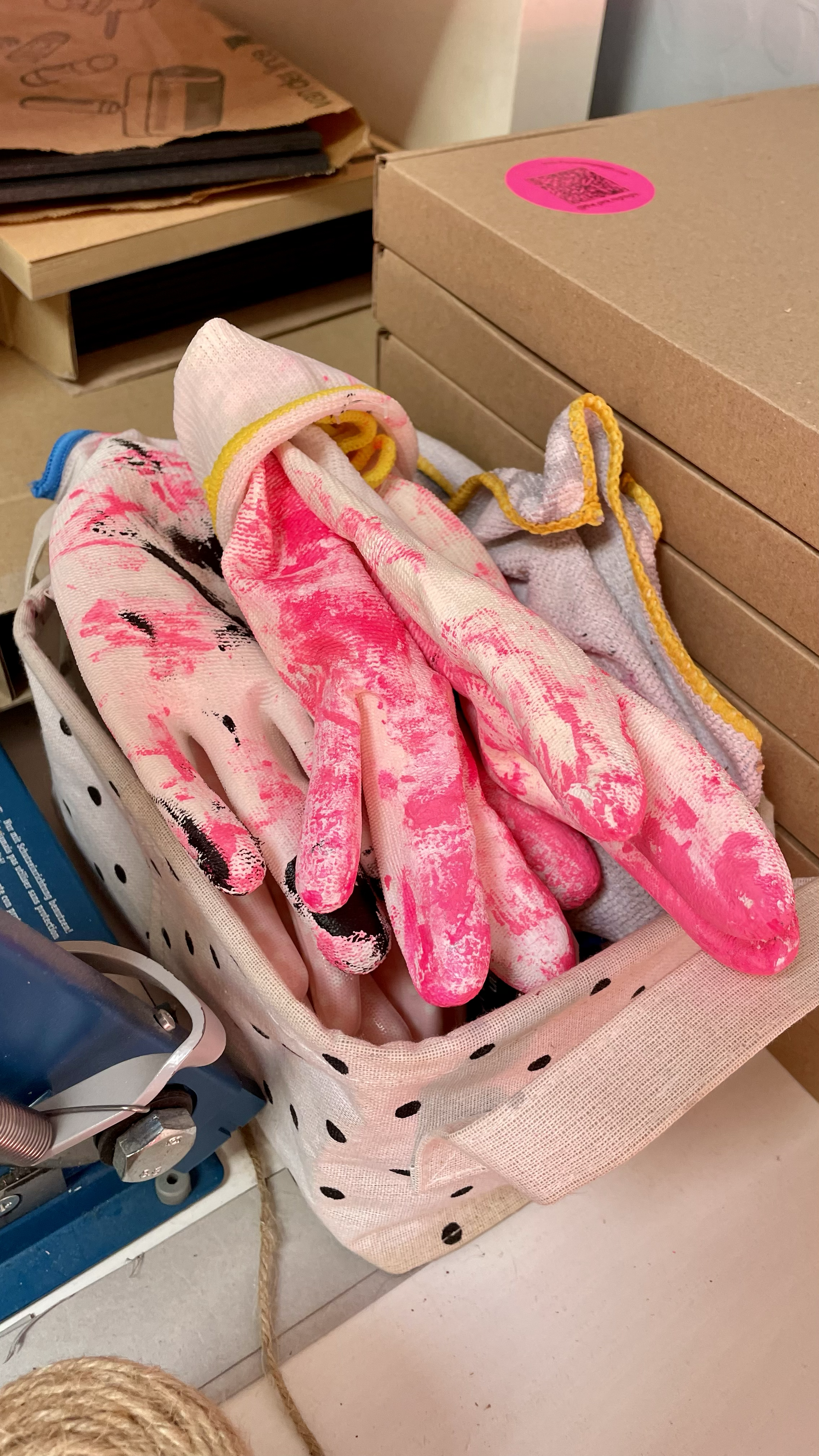 Pink and black painted gloves in a pink polka-dot basket on a shelf with cardboard boxes and paper bags in the background.
