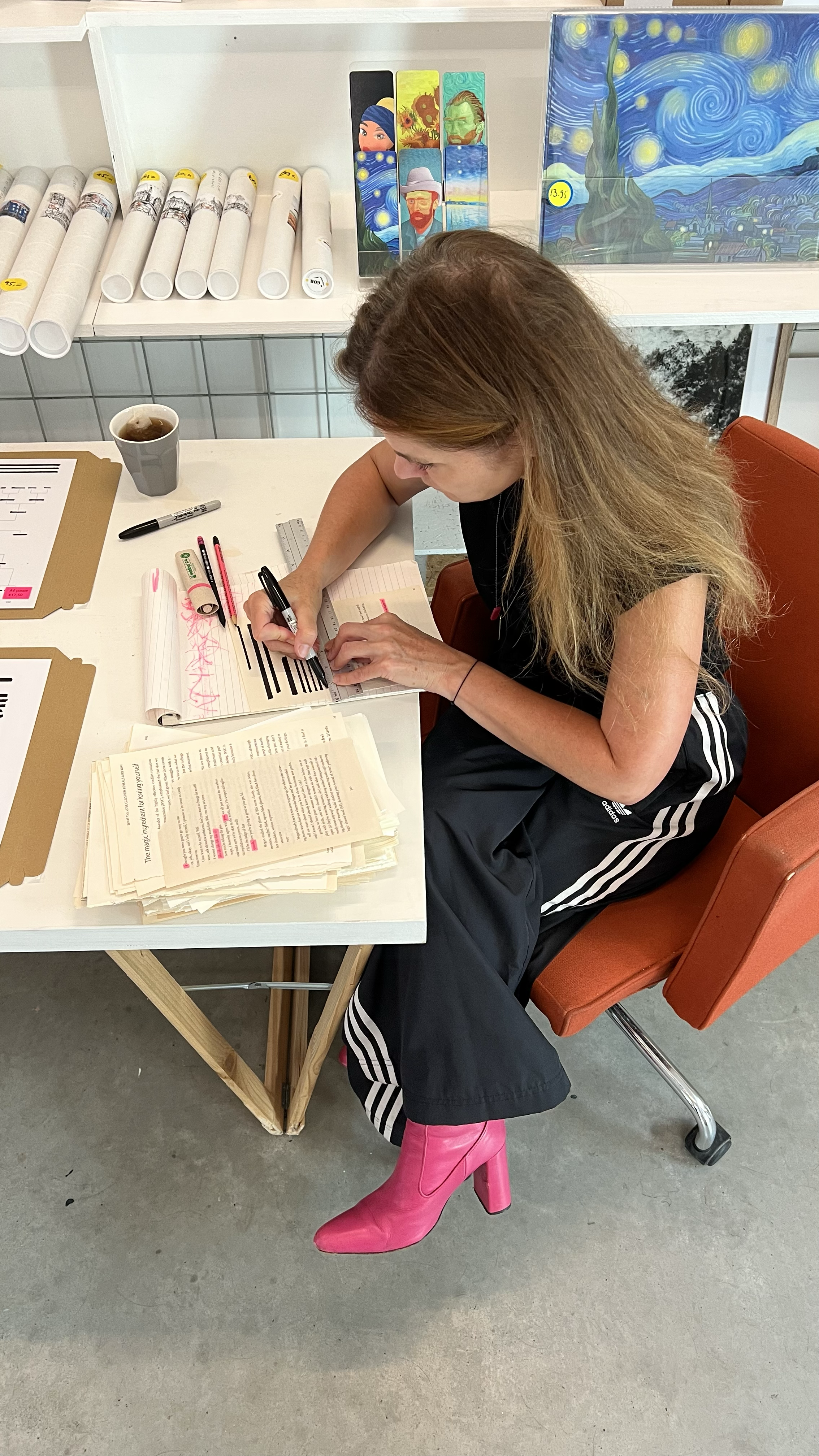 A woman with long hair wearing bright pink cowboy boots, black Adidas track pants, and a black t-shirt, sitting on an orange chair, studying papers and writing in a notebook at a white table. There are books, markers, and pens on the table, a cup of 