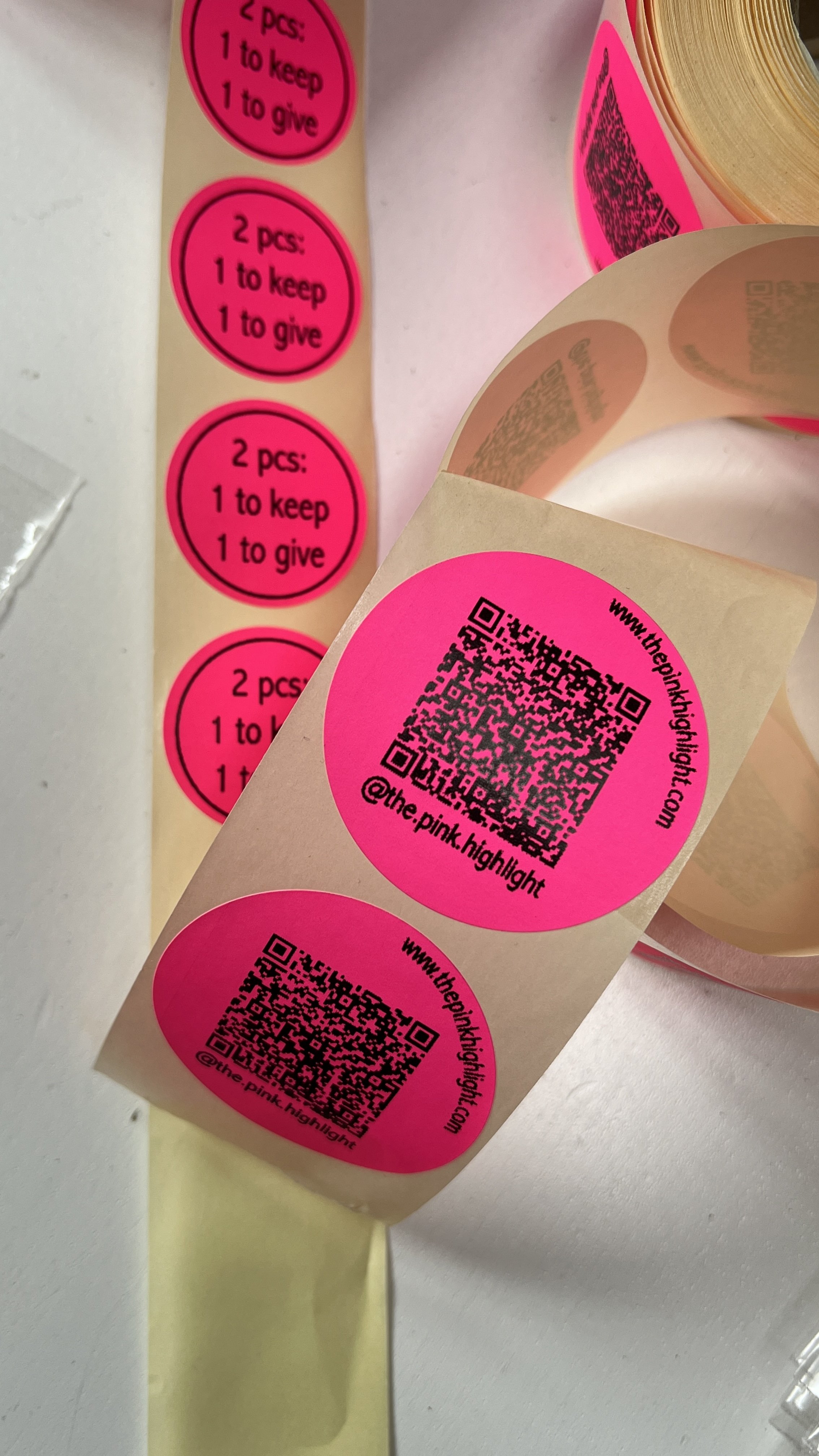 Spools of pink circular stickers with black QR codes and text, some with instructions reading '2 pcs: 1 to keep, 1 to give'.