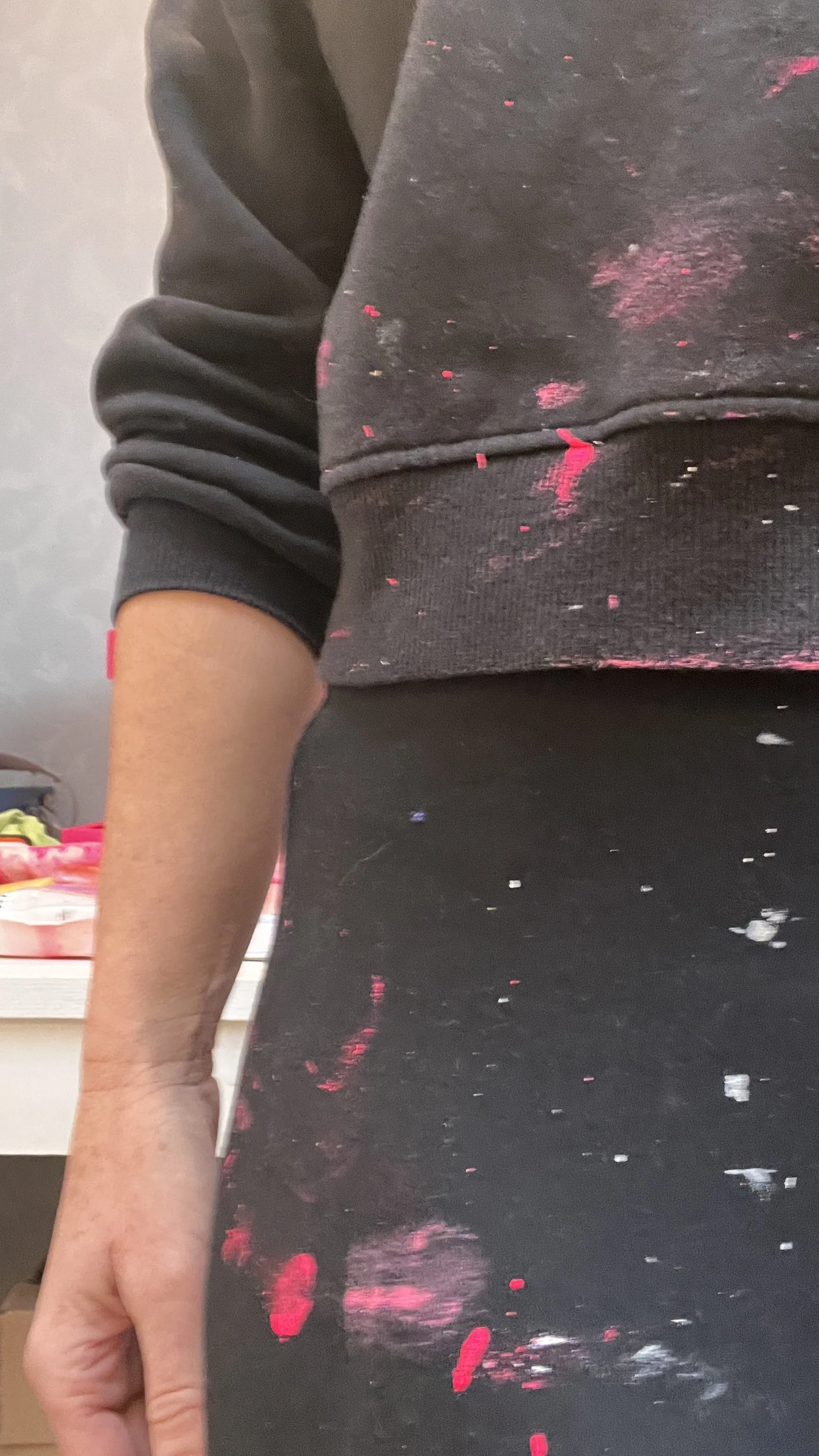 Close-up of a person wearing a black sweatshirt with pink paint splatters, standing in front of a mirror which reflects pink and white paint splatters on a dark surface.