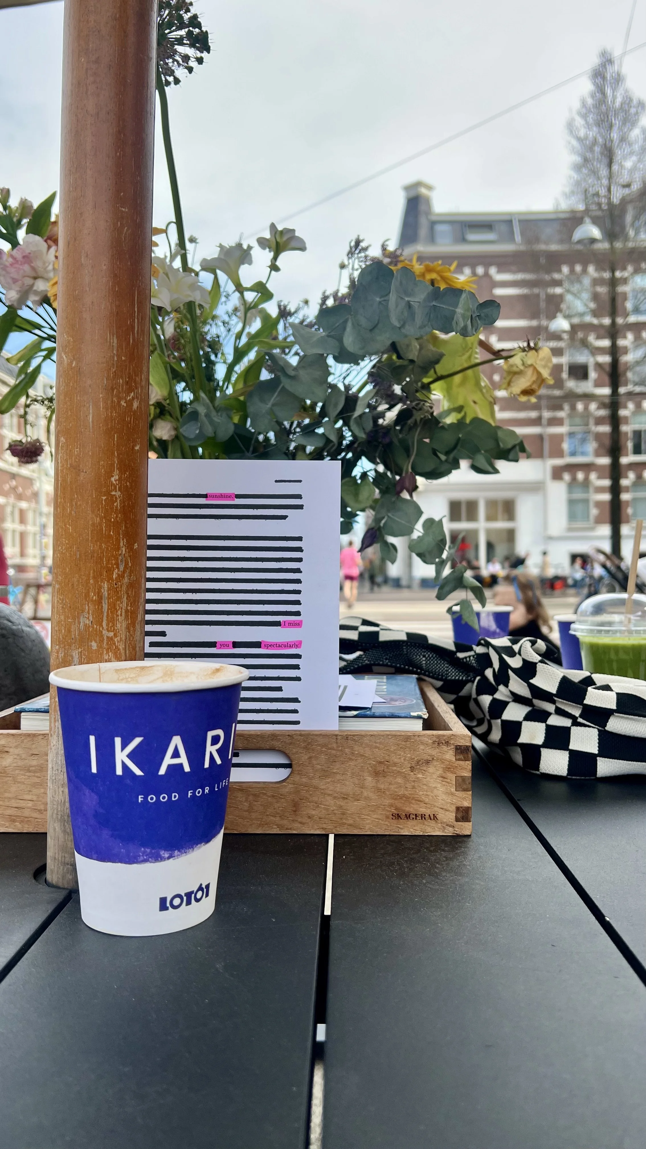 A table at an outdoor cafe with a blue and white paper cup labeled 'IKAR', a tray with a flower arrangement, a paper with pink highlighted words, and a black and white checkered bag. In the background, there are buildings and people walking.