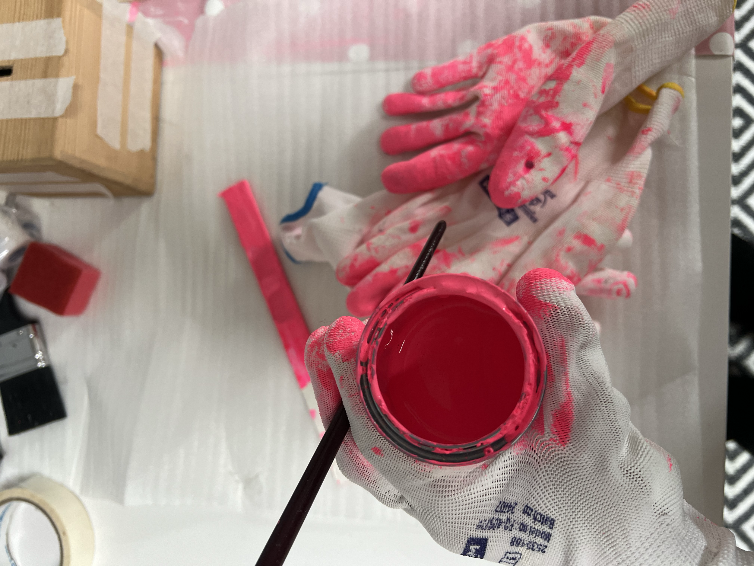 A gloved hand holding a container of pink paint with a black brush inside. The background includes a wooden block with paper and paint supplies on a white table.