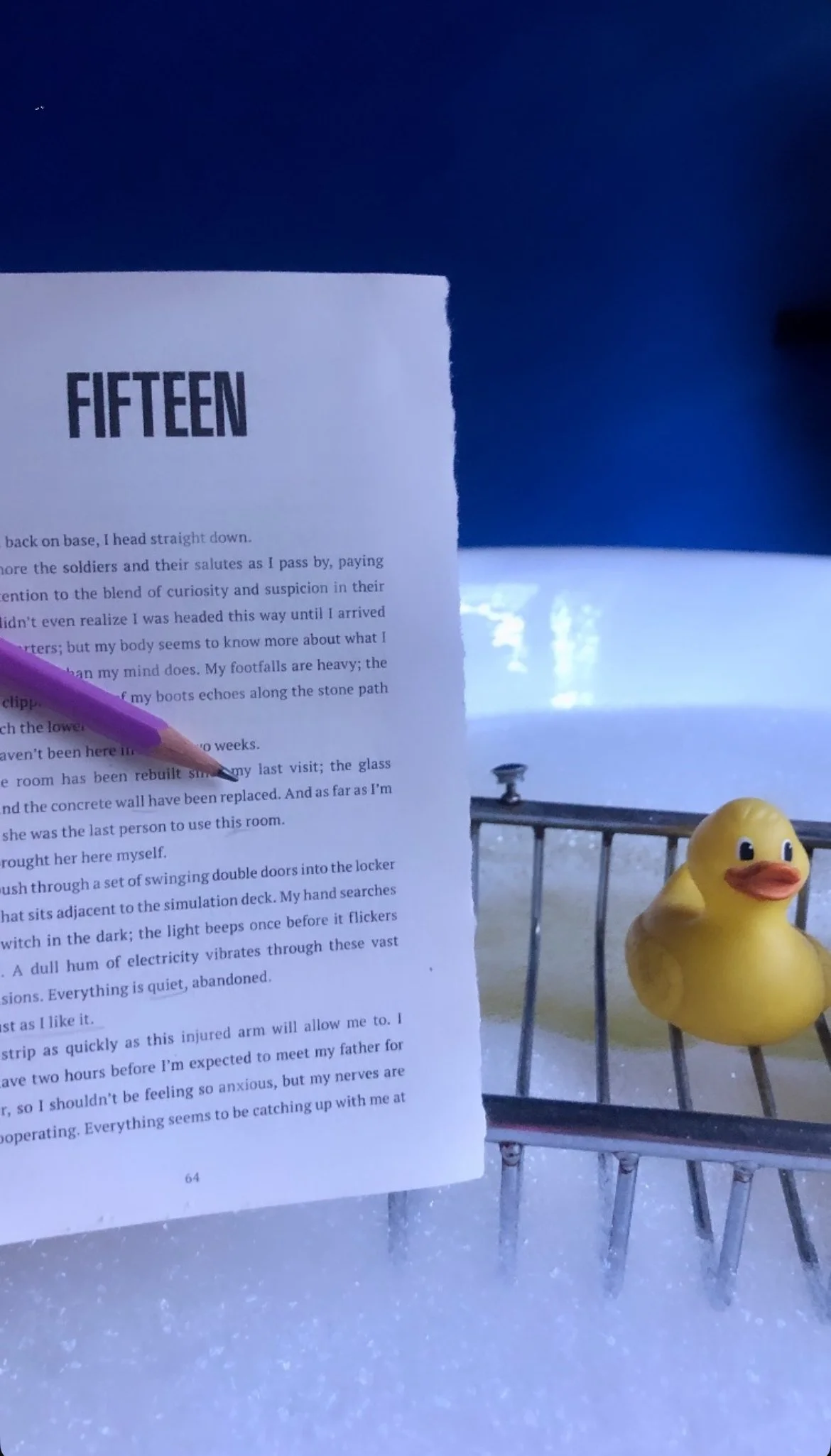 A close-up of a printed page titled 'FIFTEEN' held in front of a window showing an outdoor scene with snow, a yellow rubber duck figurine, and part of a small metal table.