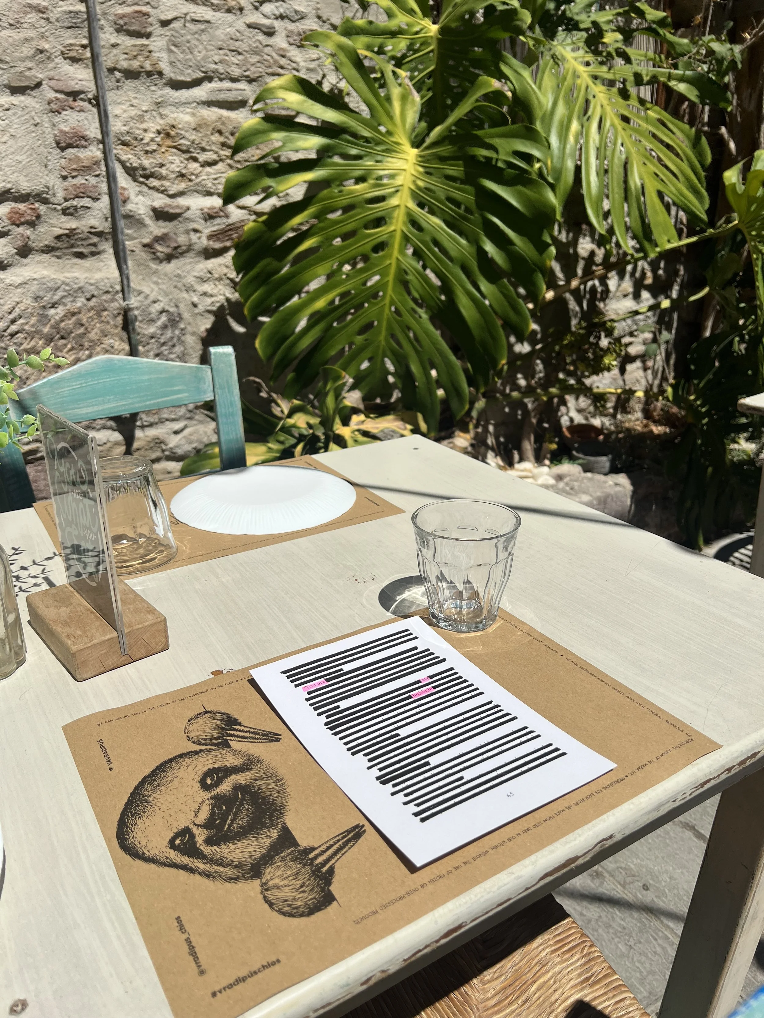 A table set for dining outdoors with a paper placemat featuring a sketch of a squirrel, a glass, and a menu card, against a backdrop of green tropical plants and a stone wall.