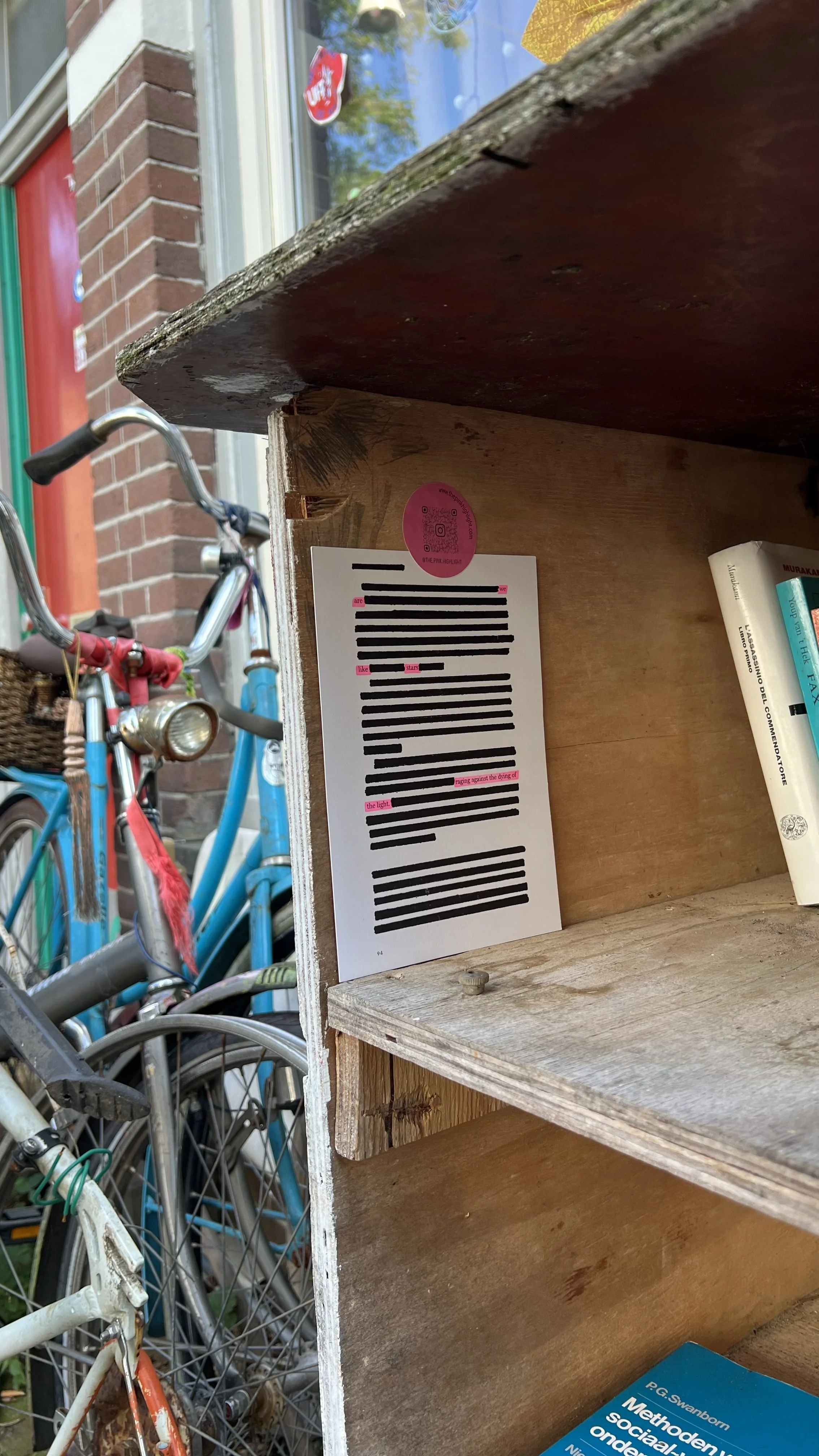 Wooden bookshelf with a flyer inserted, bicycles parked nearby, and a brick building in the background.