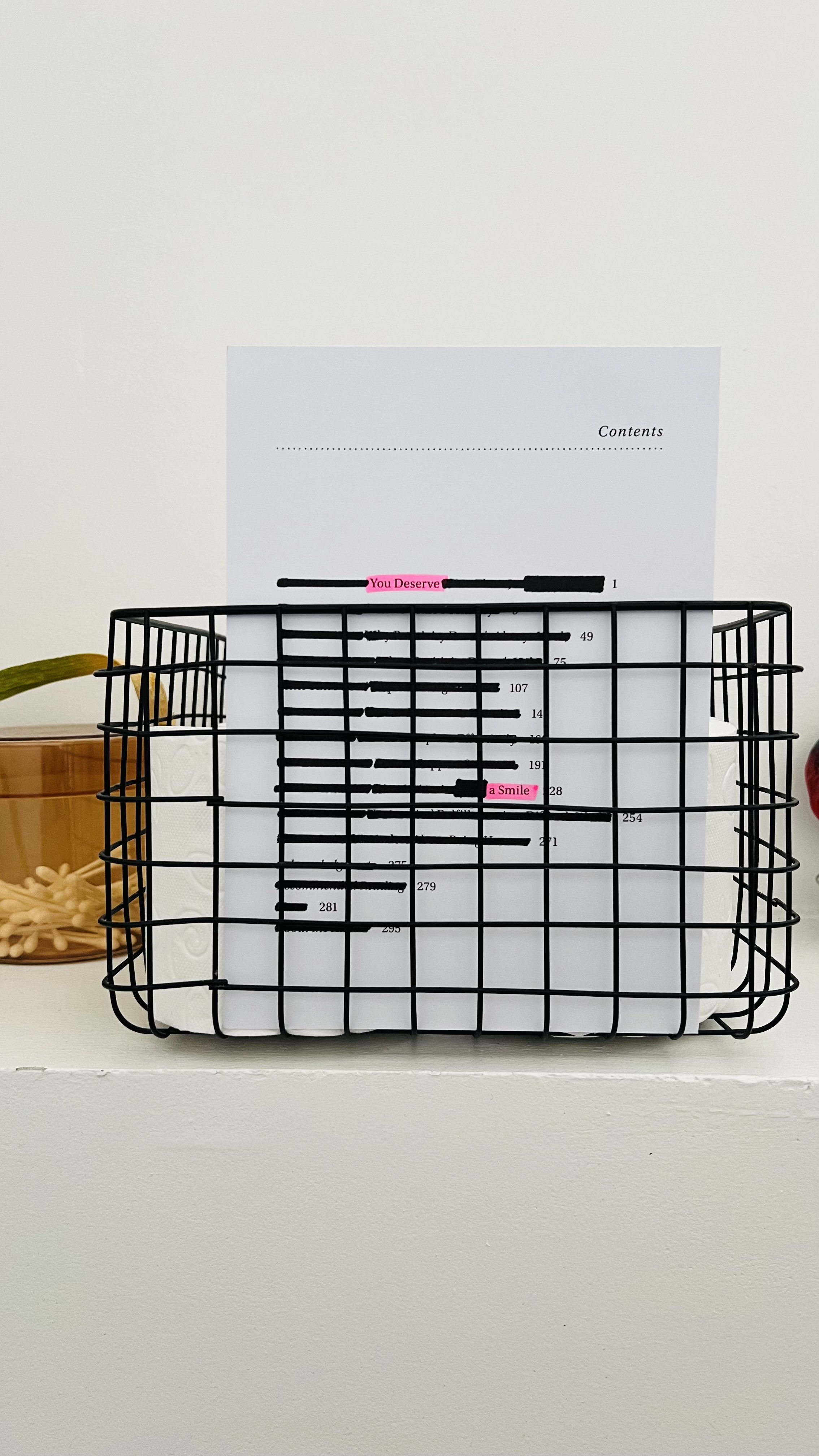 Wire basket holding a paper with the words 'You Deserve' and 'a Smile' highlighted in pink, placed on a white surface with a small plant and a container of toothpicks nearby.