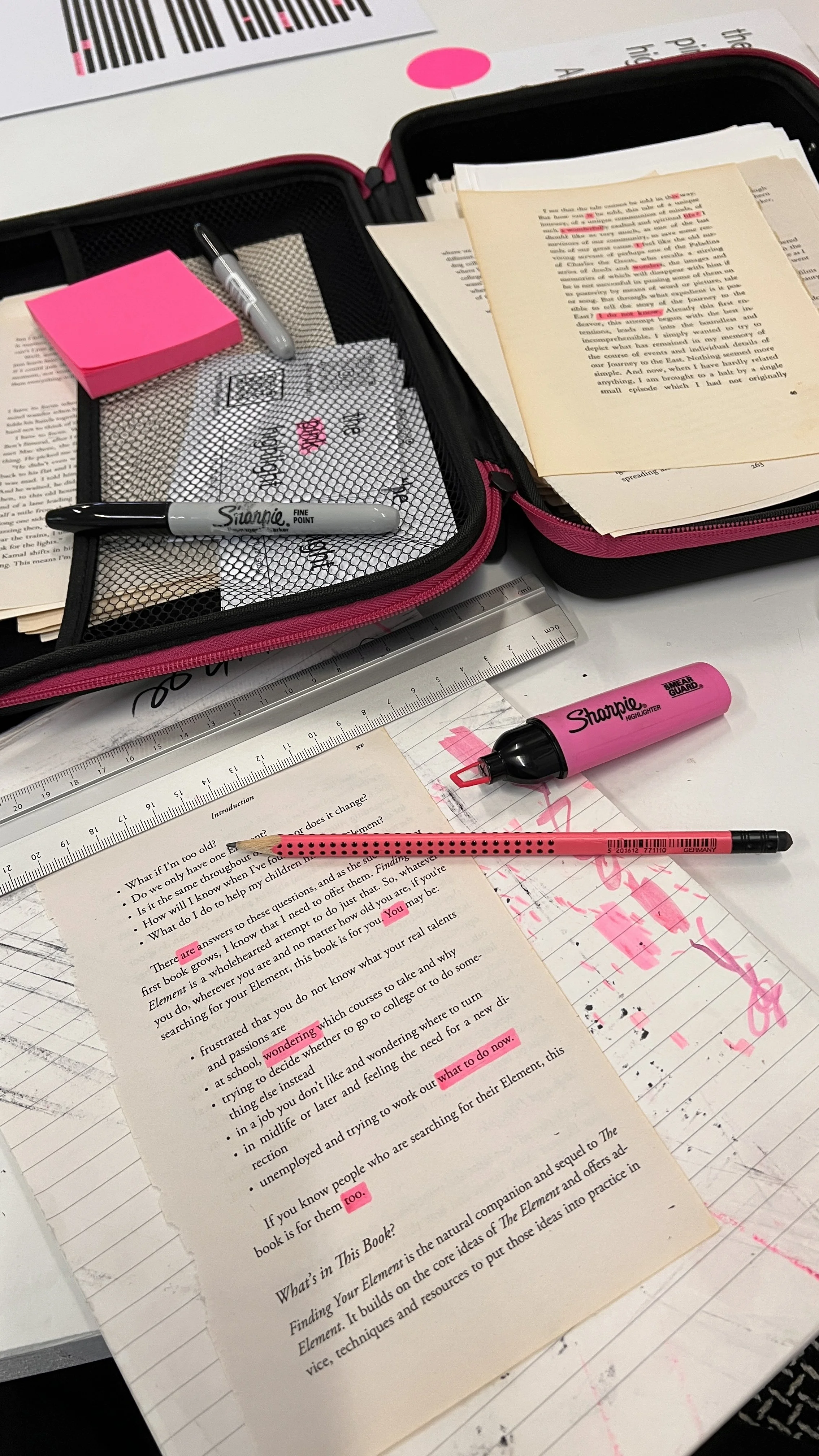 A desk with various study materials, including a book with highlighted text, a pink highlighter, a pink pen, sticky notes, a ruler, and an open pencil case containing a pen and a small pad of sticky notes.