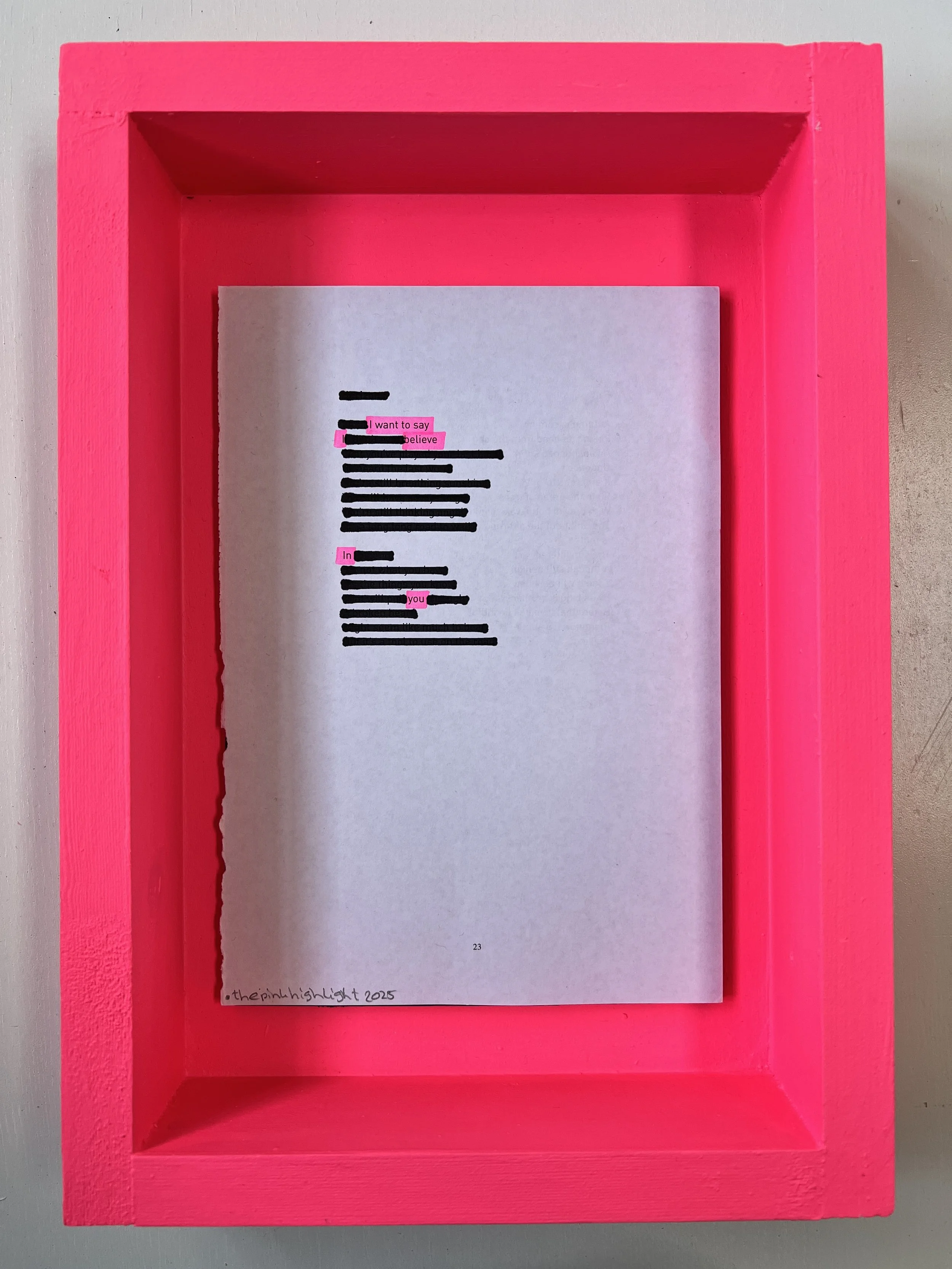 A printed sheet of paper with blacked-out text and pink highlighted words, placed inside a bright pink wooden shadow box. The sheet contains the phrase "I want to say I believe in you" with pink highlights on the words "I want to say," "believe," and