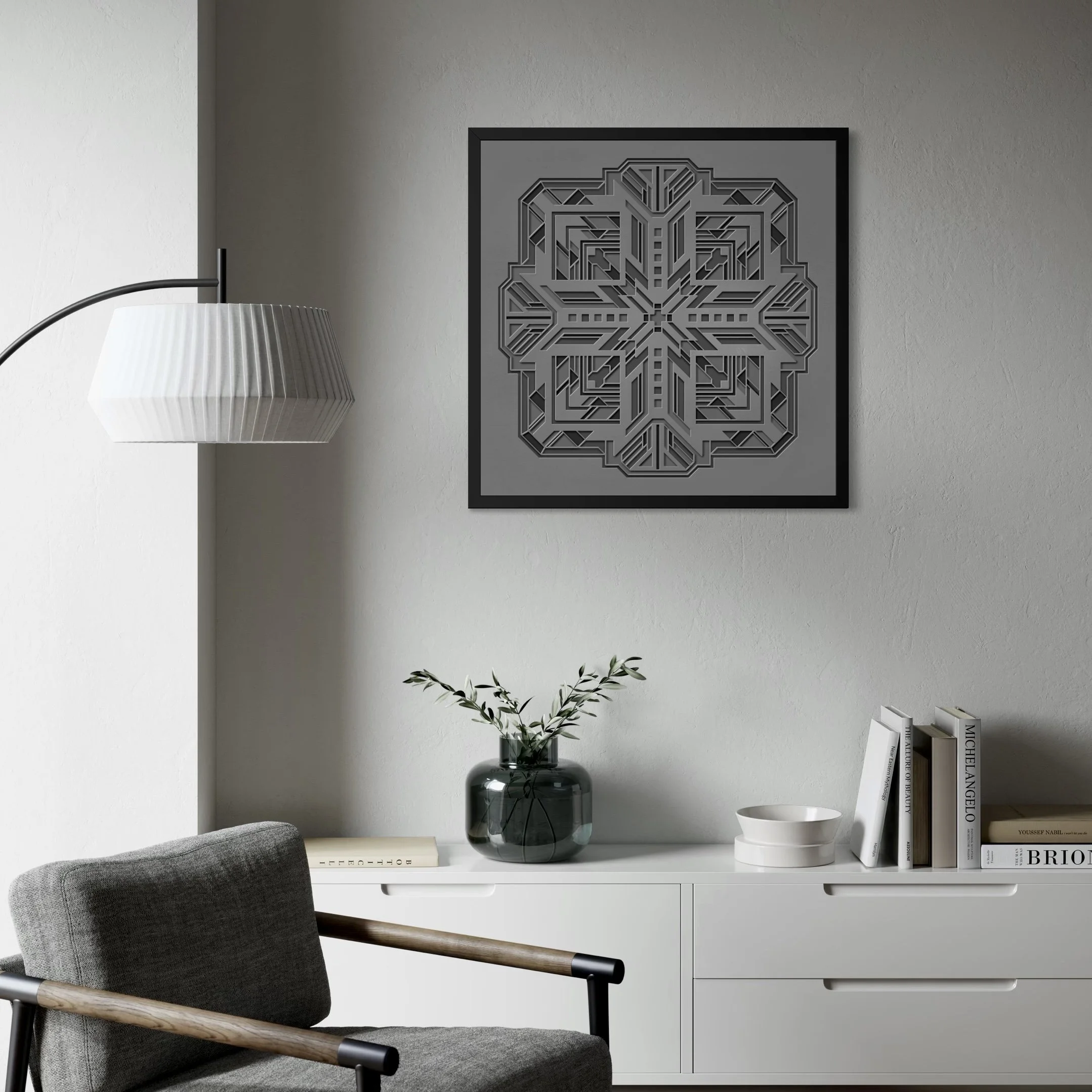 A modern home office with a white desk, grey upholstered chair, monochrome geometric art piece on the wall, a white pleated lampshade, a glass vase with plant, white books, and white ceramic bowls.