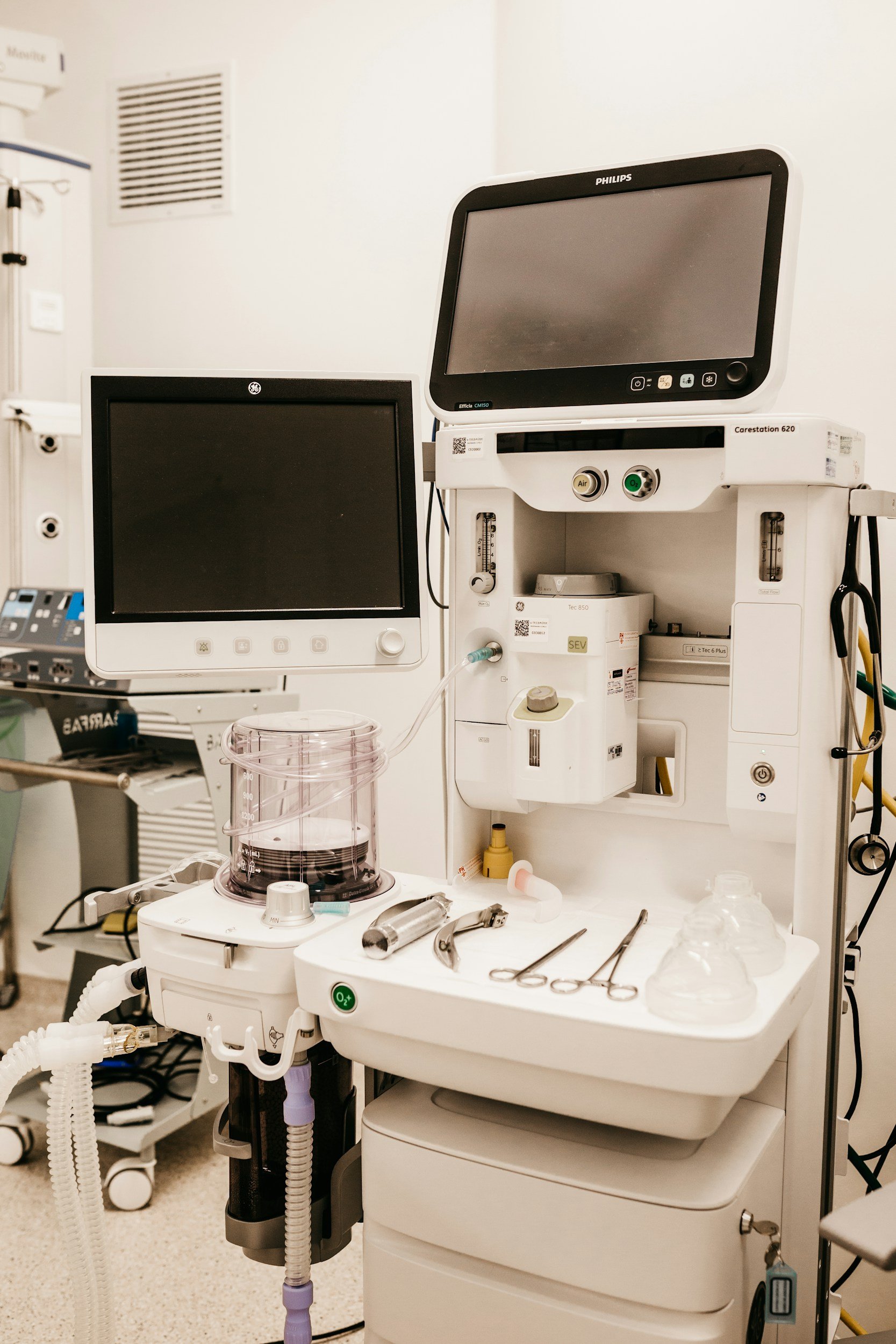 Hospital operating room with multiple medical monitors, surgical instruments, and anesthesia equipment.