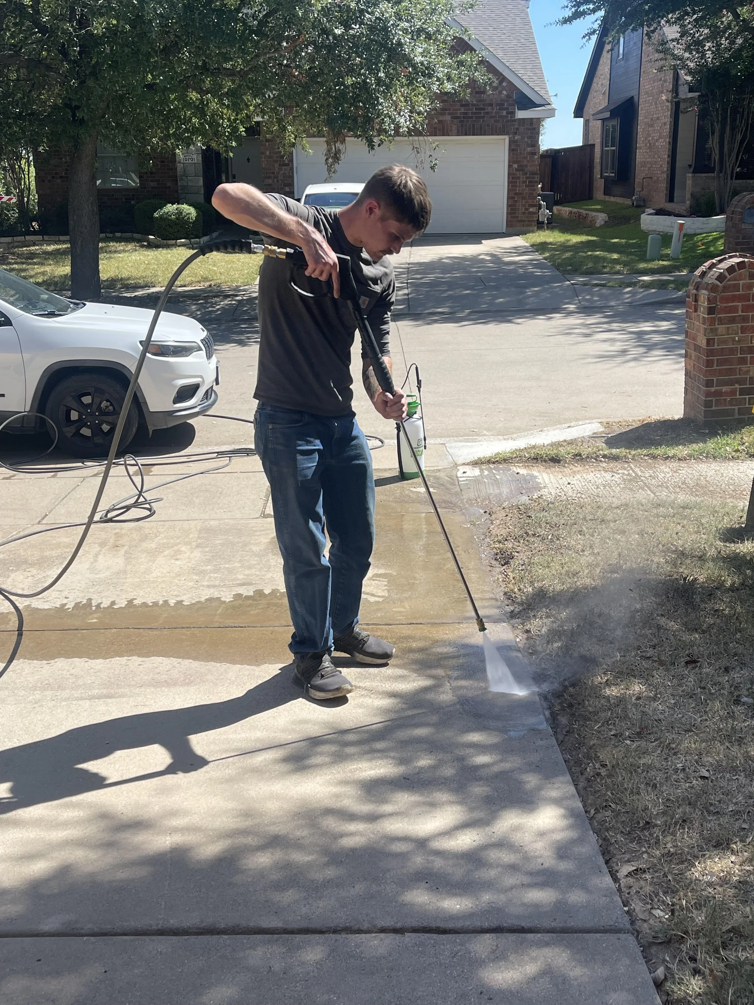 A man is pressure washing a concrete driveway on a sunny day in front of houses. He is wearing a black long-sleeve shirt, jeans, and grey shoes, holding a pressure washer wand aimed at the driveway, with water spraying out. There are trees, a white car parked on the street, and houses in the background.