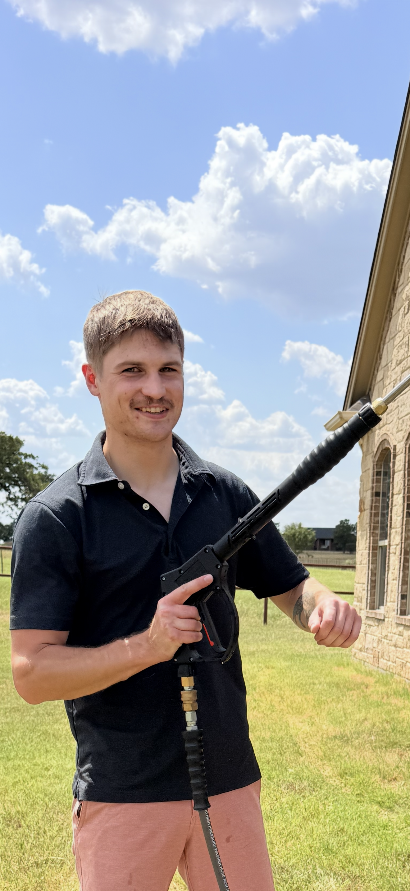 A young man standing outside, smiling, holding a power washer with a long nozzle directed upwards. He is wearing a black collared shirt and pinkish shorts, with a tattoo on his left arm. The background shows a grassy yard, a brick house with stone accents, a clear blue sky with scattered clouds, and some distant trees and structures.