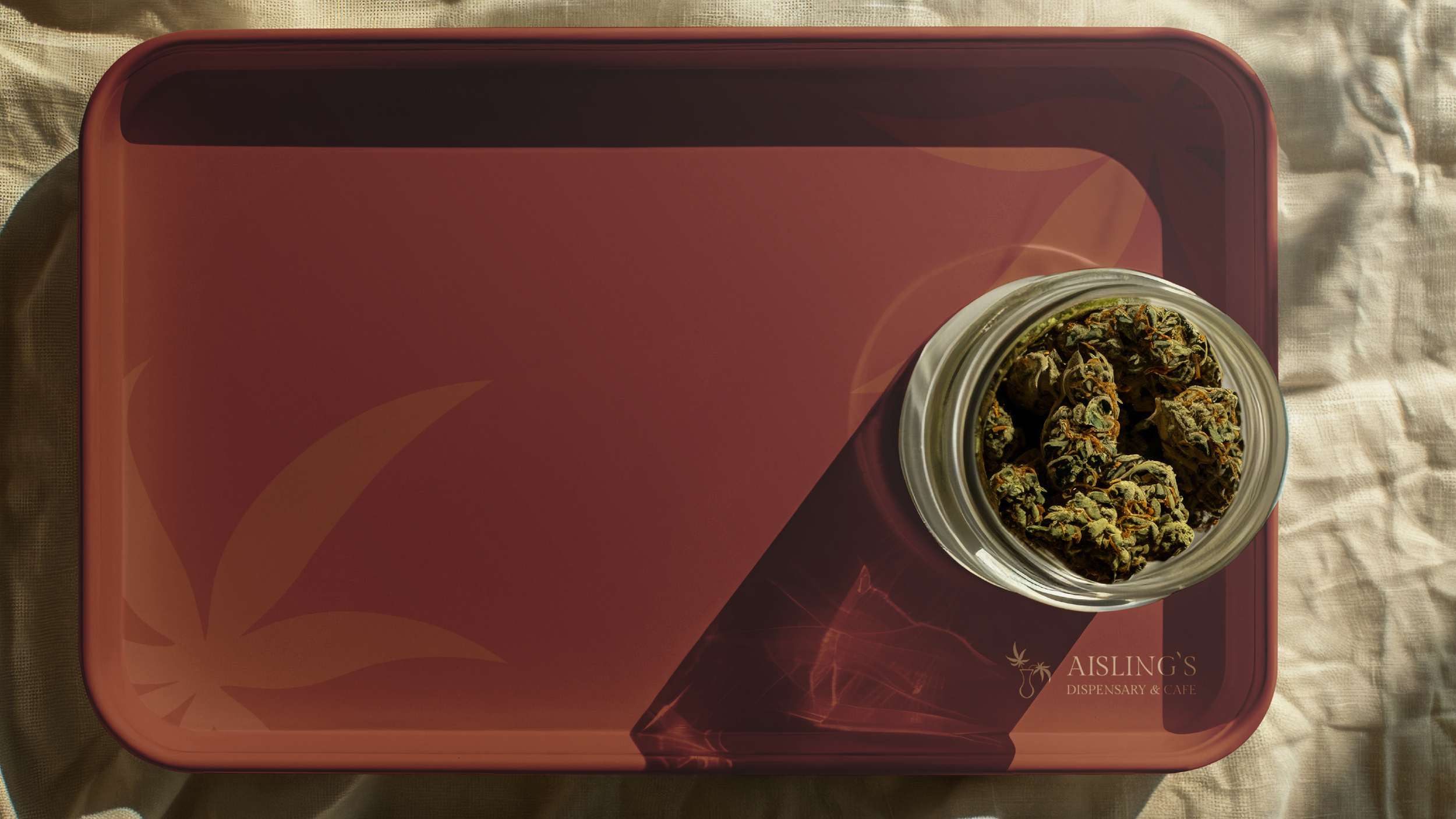 A glass jar filled with dried cannabis buds placed on an Aisling's branded maroon tray. The tray is on a beige fabric surface.