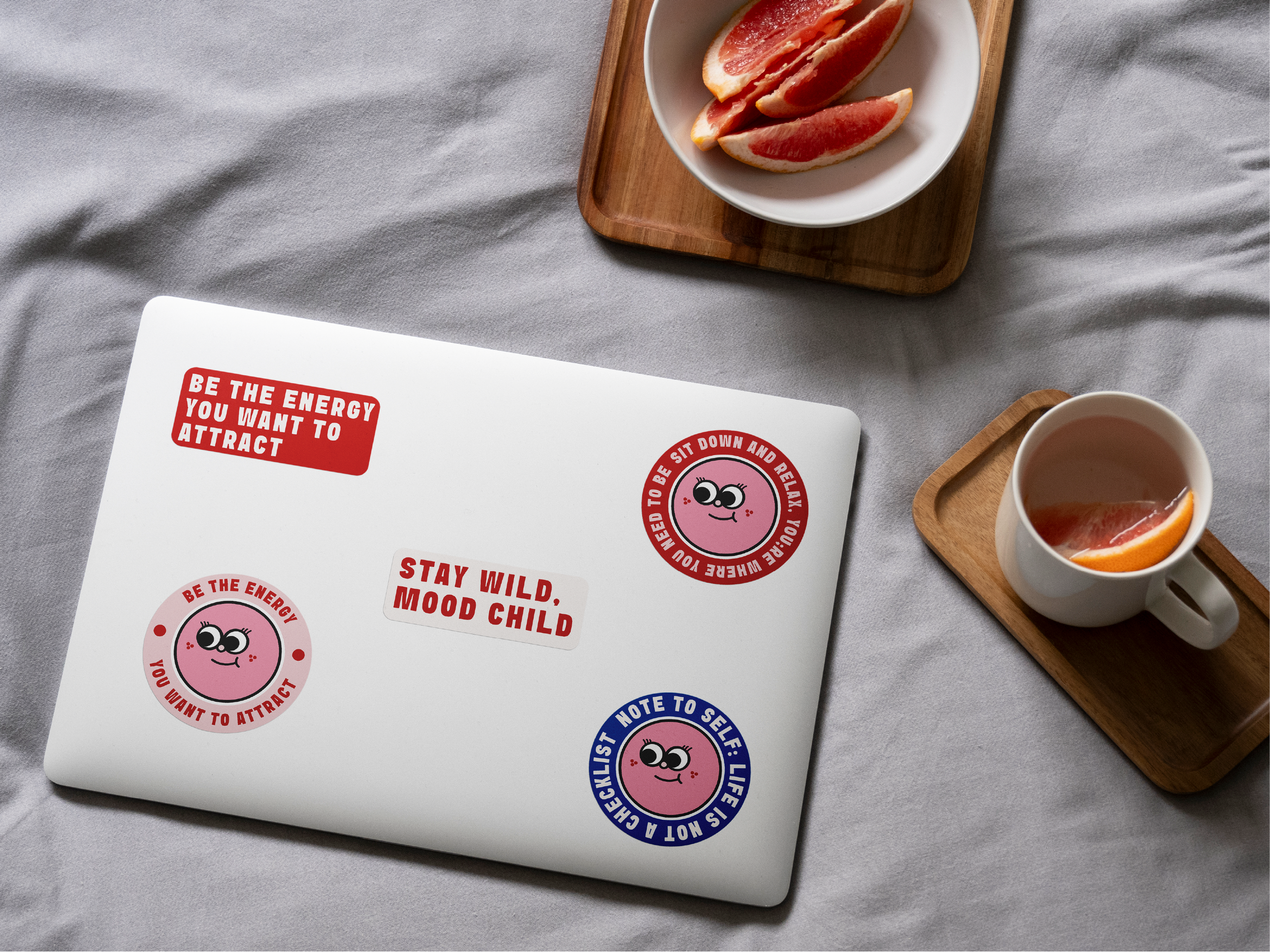 A closed laptop with red, pink, and blue Soul Circle stickers on top, next to a white bowl with grapefruit slices on a wooden tray and a white mug with hot beverage on a gray fabric surface.