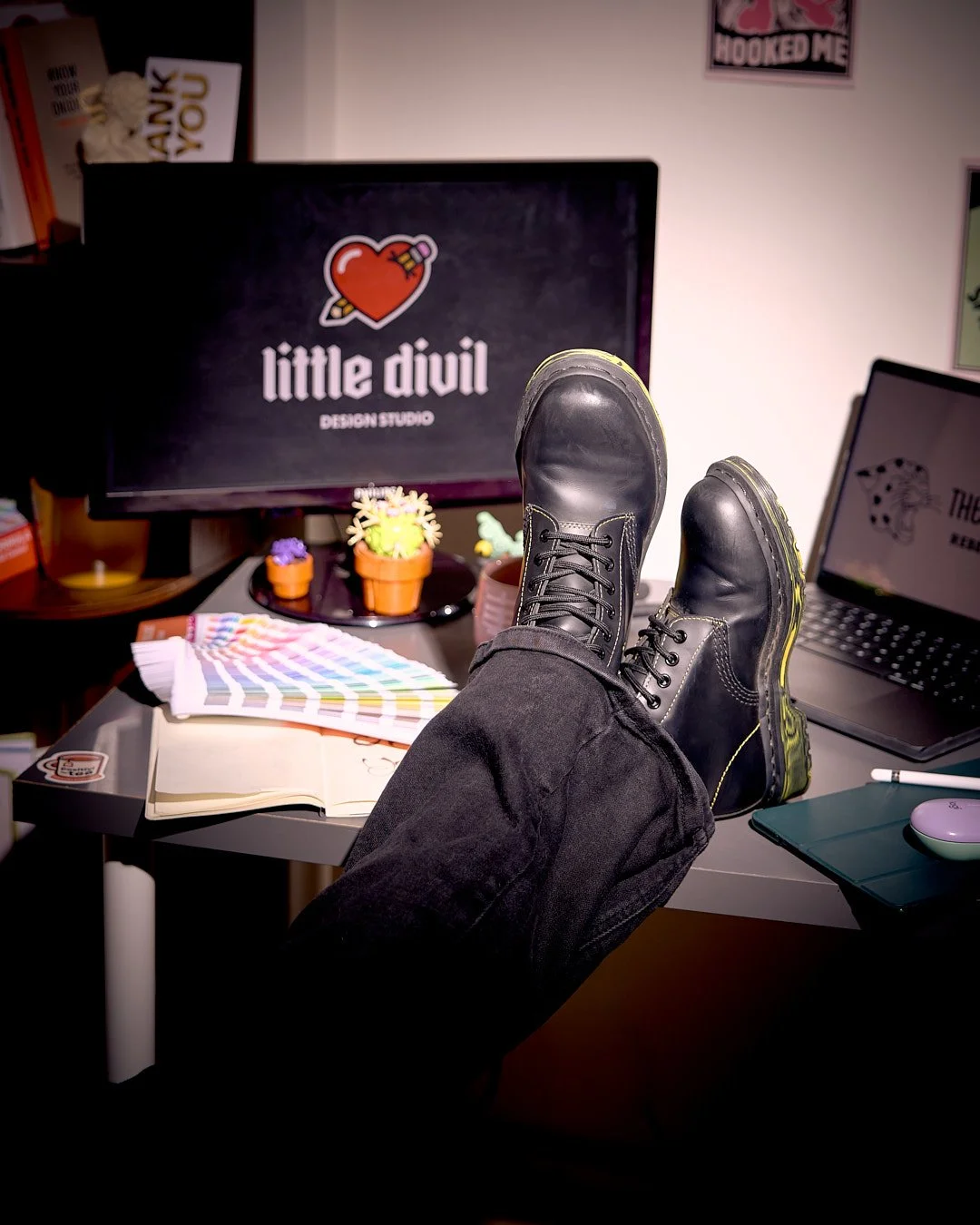 Amy's black dr Martens propped up on the desk, computer screens displaying design work, color swatches, and decorative cacti on the desk. The background shows a monitor with a "little divil" logo and a wall with posters.