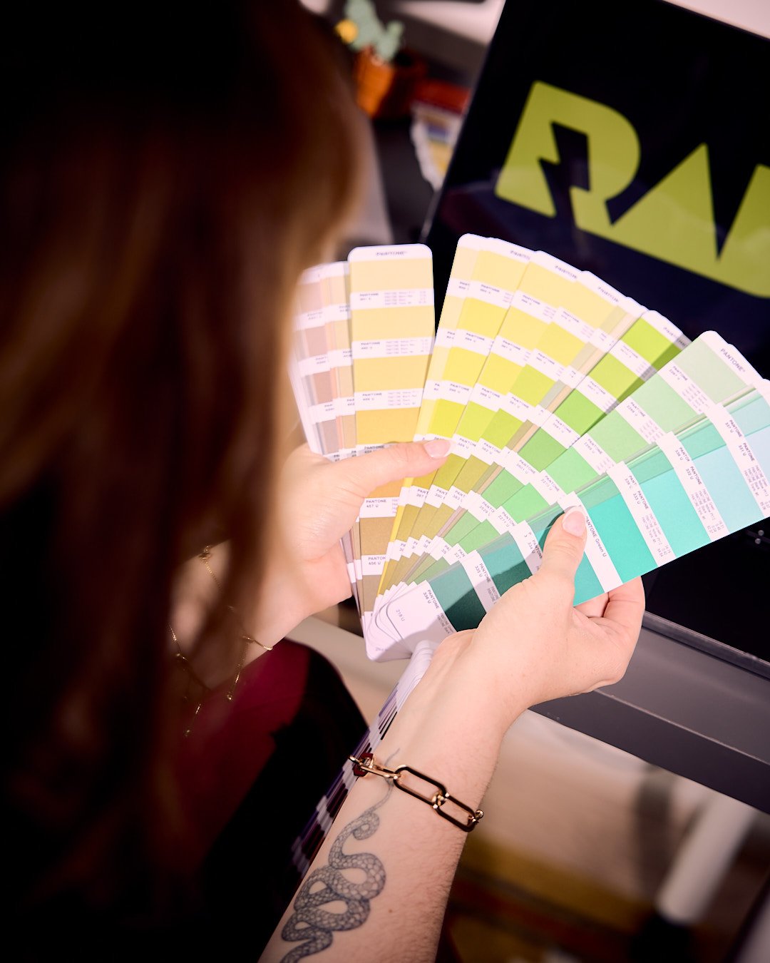 Person holding a fan deck of Pantone colour swatches with shades of yellow, green, and blue, in front of a laptop displaying a logo.