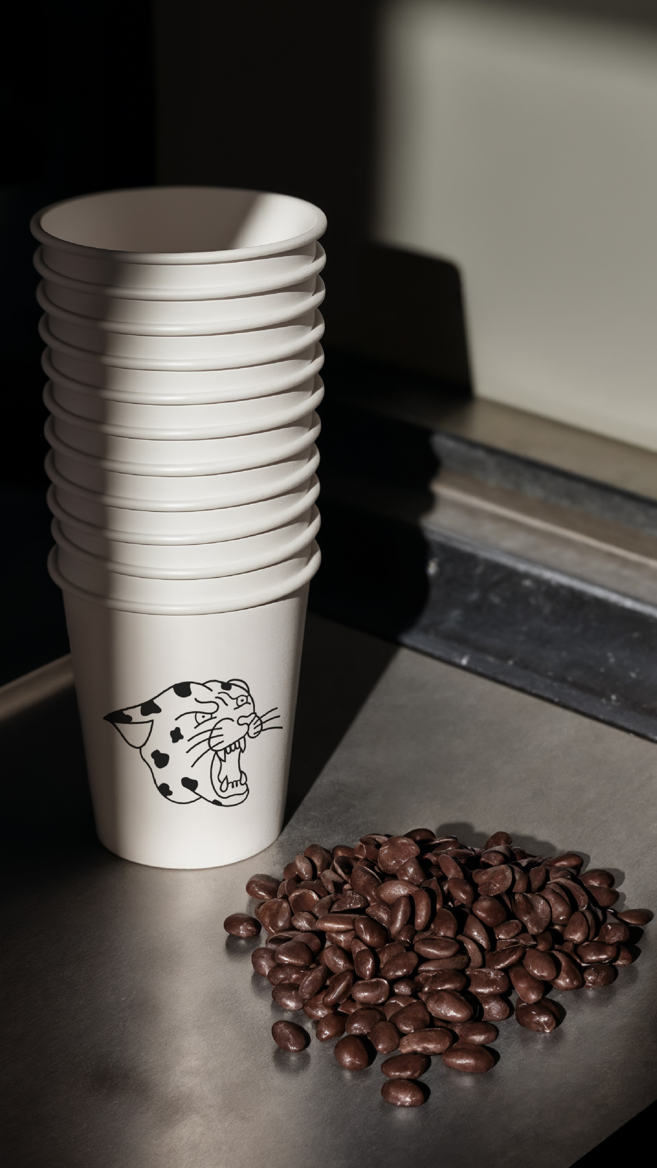 Stack of white disposable cups with The Daily Grind logo on the front, placed on a countertop with a pile of coffee beans in a coffee shop or café setting.