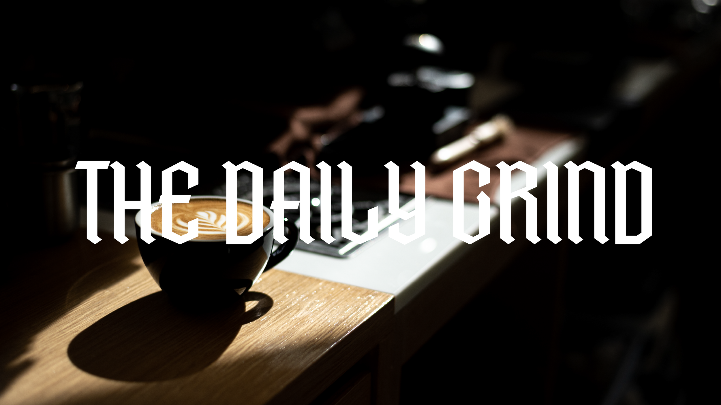 Close-up of a latte in a cup with latte art, on a wooden table with a keyboard and a hand in the background, with the words 'THE DAILY GRIND' overlaid.