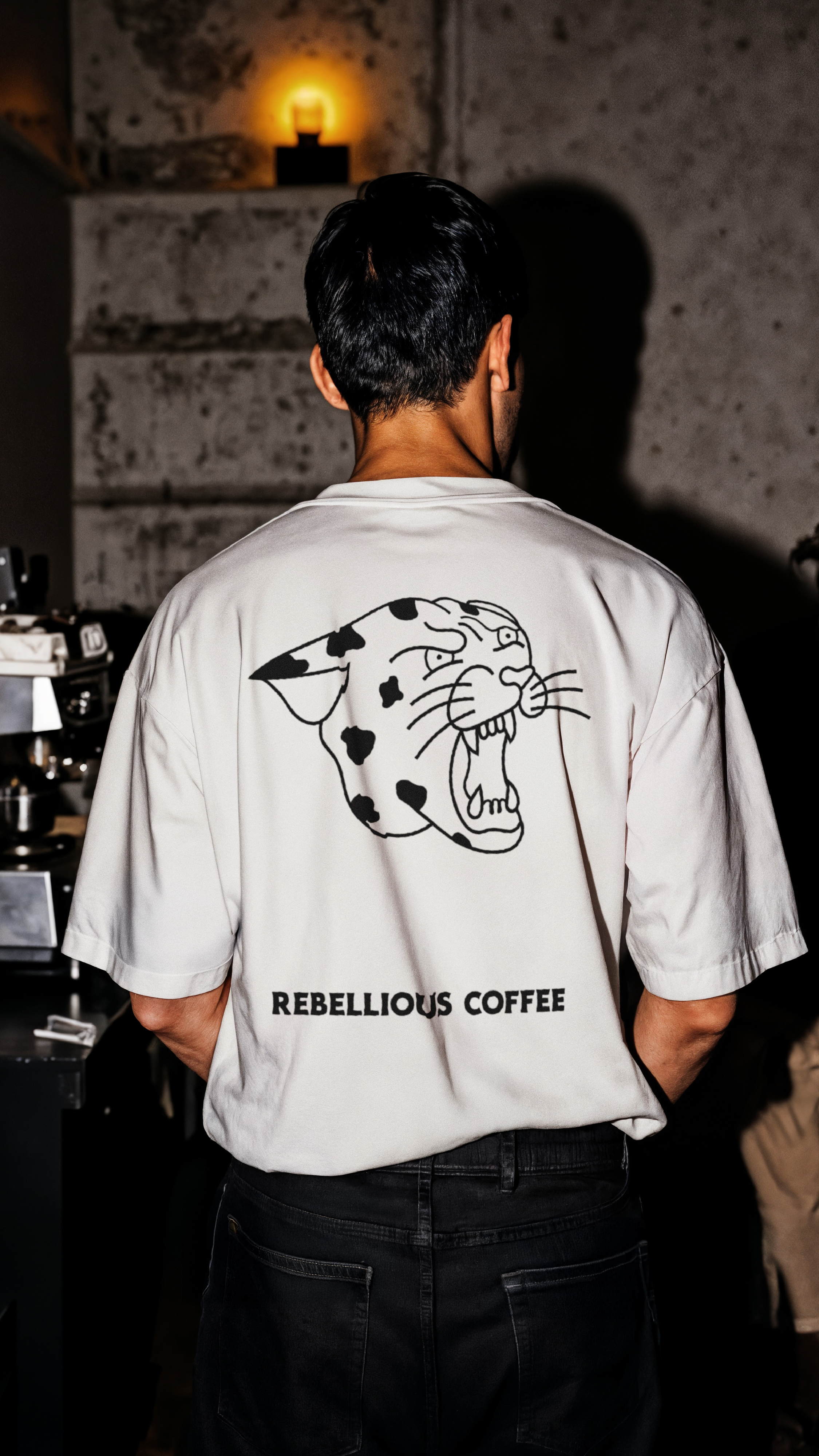 Barista wearing a white shirt with a cheetah graphic and the words 'Rebellious Coffee' standing in a dimly lit indoor space with a warm yellow light in the background. They are facing away from the camera.