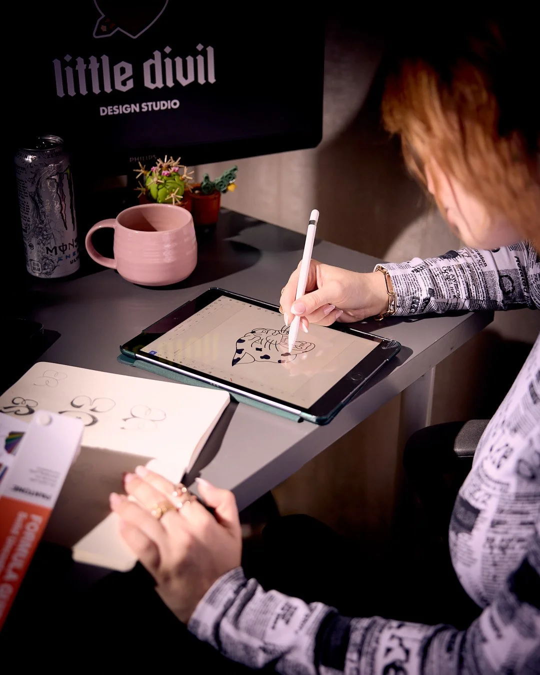 Person illustrating a cheetah on a tablet with a stylus, sitting at a desk with a sketchbook, a pink mug, a soda can, and small cactus decorations.
