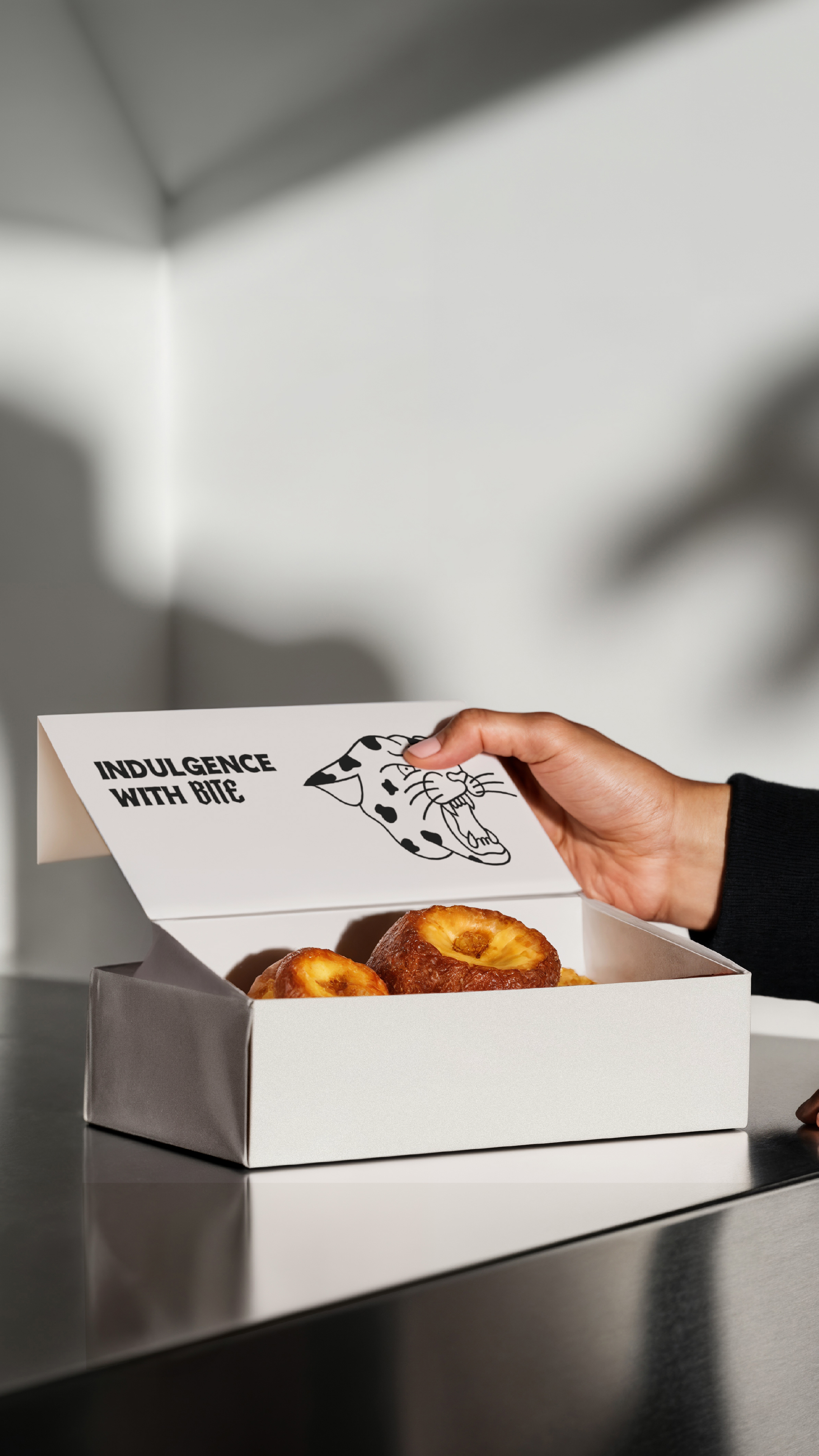 Hand opening a white box of donuts with a label that says "Indulgence With Bite" and displays The Daily Grind logo.