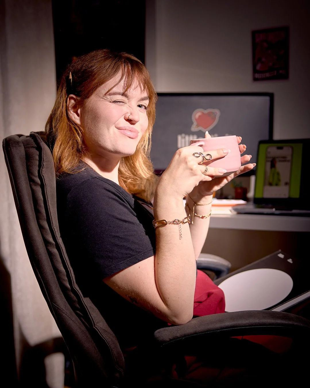 A woman with red hair holding a pink mug, winking at the camera in an office setting.