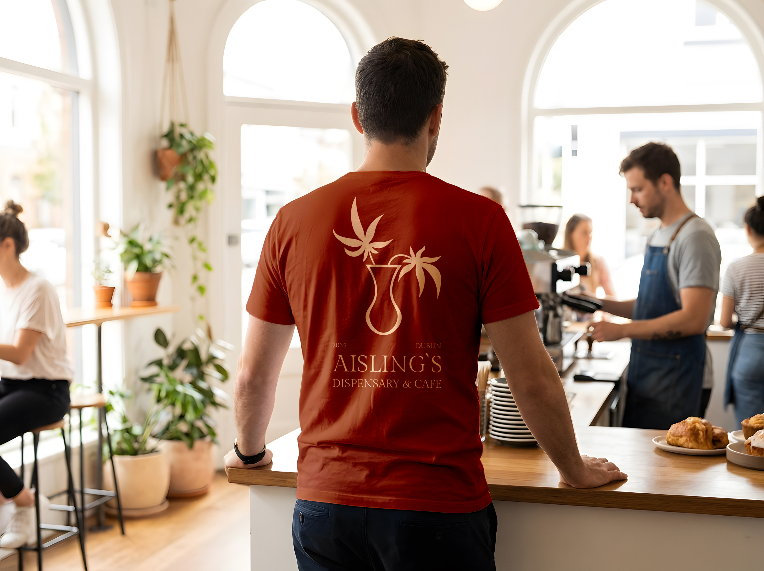 A waiter wearing an Aisling's branded t-shirt featuring the cannabis leaf logo on the back, standing at a cafe counter with other people around, and sunlight coming through large windows.