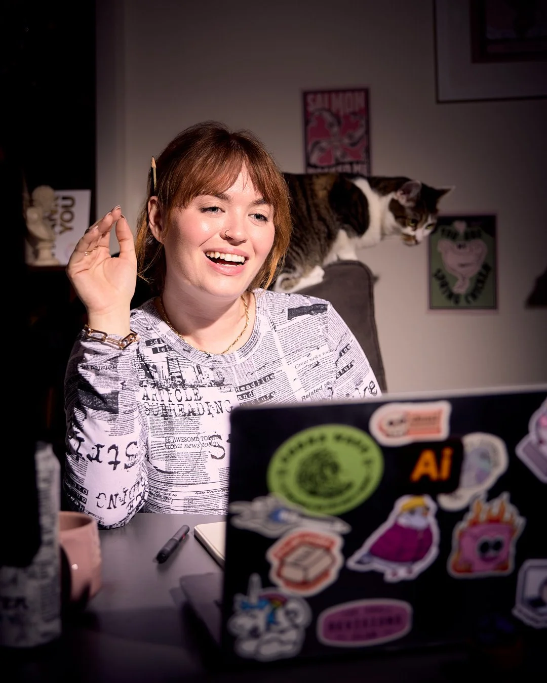 A smiling woman with short ginger hair, wearing a newspaper print shirt, sitting at a desk with a laptop covered in stickers, a pink mug, a pen, and a notebook. A cat is perched on the back of her chair behind her.
